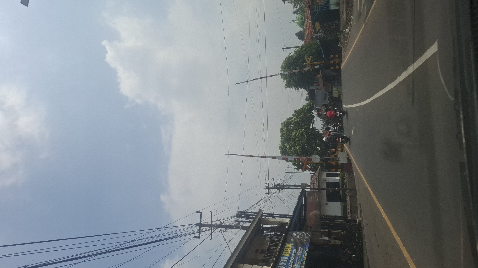 The image depicts a street scene in Sukasari, Indonesia, characterized by a railway crossing. In the foreground, a paved road with yellow and white lane markings curves into the distance. Several motorcycles are visible on the road, with riders on them, suggesting ongoing traffic. To the left, a building with a sign that appears to read "ARYA BAN" is situated alongside the road, with a display of tires and possibly a motorcycle parked in front. Utility poles with numerous overhead wires dominate the left side of the frame.
The central focus of the scene is the railway crossing, marked by two red and white striped barrier gates, one on each side of the road, which are raised. A train track is implied by the presence of these gates and a red and white signal light. Trees, including a large, dense one, are present behind the crossing. In the background, other buildings and structures can be seen, including what appears to be a green-domed building, possibly a mosque, on the right. The sky is partly cloudy, with a clear blue expanse interspersed with white clouds, indicating daytime conditions. The overall impression is one of a typical street in a populated area with active transportation infrastructure.