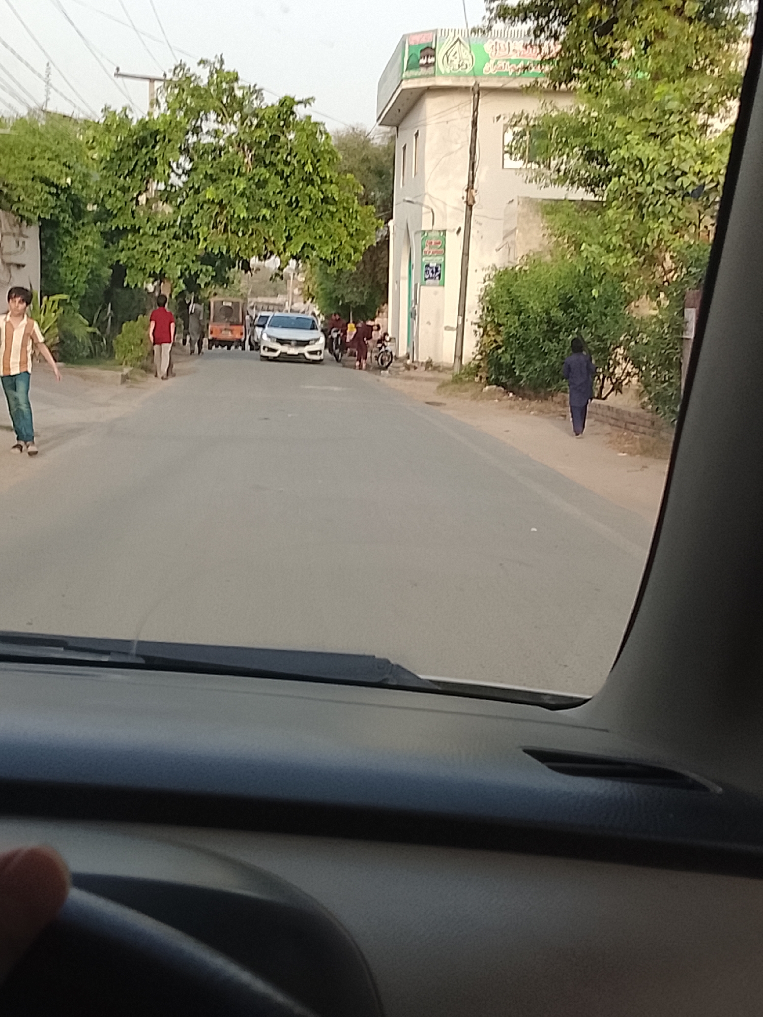 Captured from inside a vehicle, the image shows a street in Nasheman Colony, Pakistan. The road leads towards a white car ahead. A man walks on the left sidewalk, and another person in a red shirt walks on the road. A building with Arabic script on top stands on the right. A person in dark clothing walks on the right sidewalk. The image is taken from the driver's seat, with the dashboard and part of the steering wheel visible. Green foliage is prominent on both sides of the street. A auto rickshaw is visible in the road.