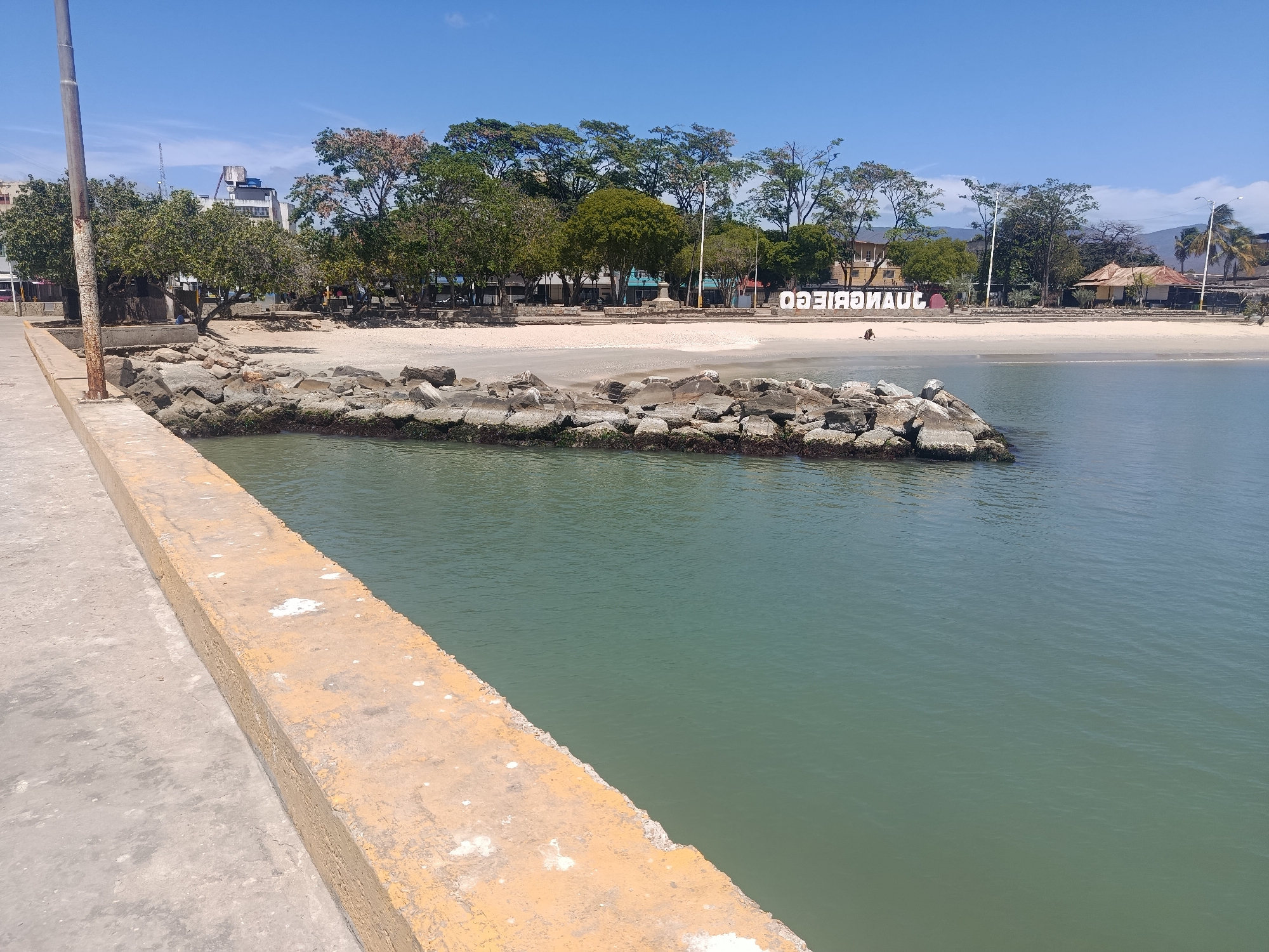 The image depicts a waterfront view in Juan Griego, Venezuela, on a clear, sunny day. A concrete pier with a yellow-painted edge is visible in the foreground. A breakwater of large rocks stretches into the greenish-blue water. Beyond the breakwater, there is a sandy beach with trees along the shoreline. In the background, there is a white sign spelling out "JUANGRIEGO" in reverse order, suggesting that the photo was taken from the water side, facing toward land. There is some sparse vegetation and building structures towards the upper left of the image. One person, or an object resembling a person, can be seen on the sand, near the water's edge.