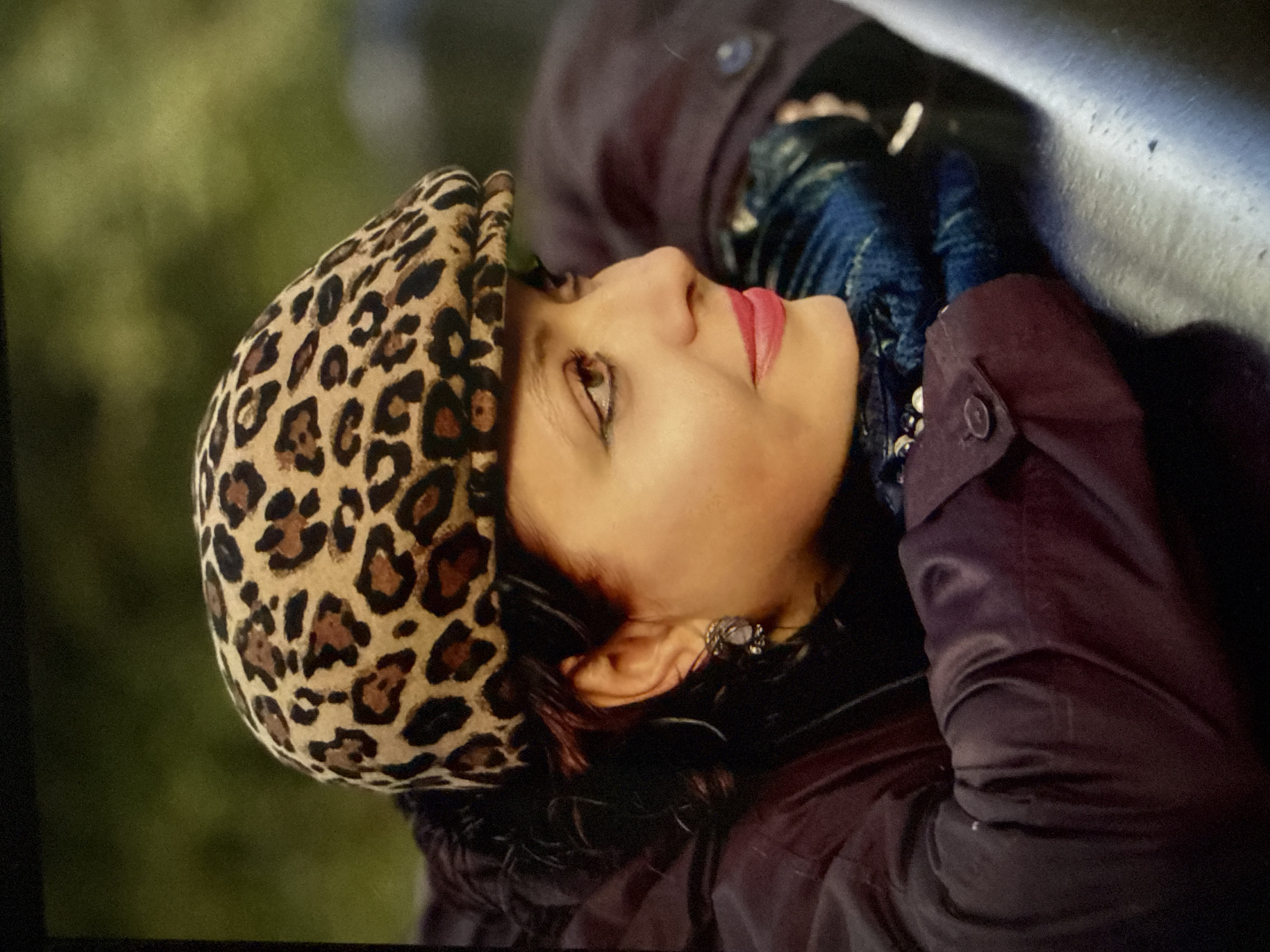The image is a portrait of a woman from the chest up, positioned in a profile view facing right. She is wearing a leopard-print flat cap, a dark jacket, and blue denim gloves. Her head is tilted down, resting on her hands which are clasped in front of her. She has dark hair and is wearing noticeable eye makeup and bright pink lipstick. She has a dangling earring in her left ear. The background appears to be a blurry outdoor setting with green foliage. There is no indication of Khabarovsk, Russia, or any specific city.
