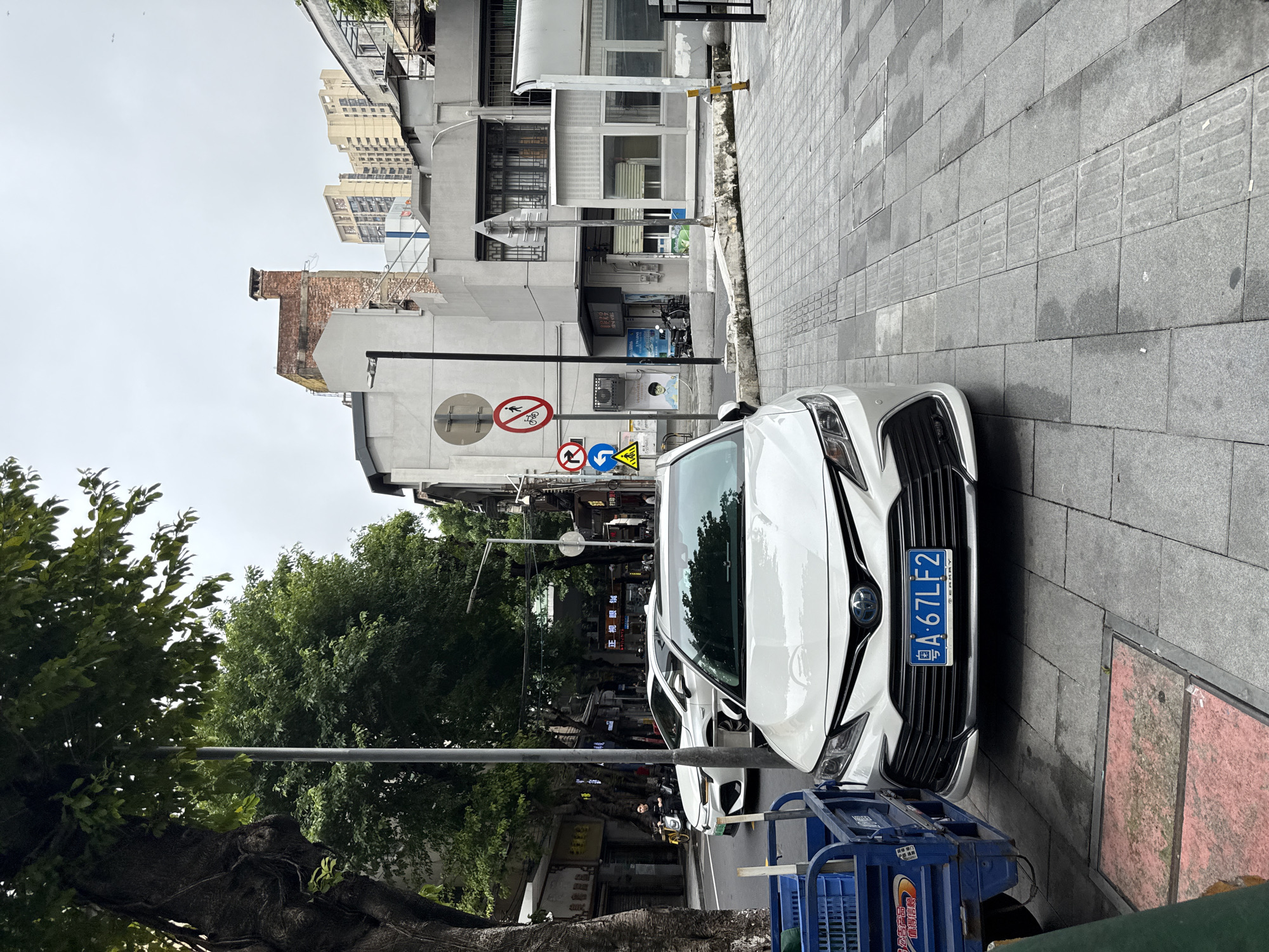 The image depicts a street scene in Liwan District, China. In the foreground, a white Toyota Camry with a blue license plate reading "A 67LF2" is parked on the right side of the road. To the left of the car, a blue wheeled cart with a green crate is visible. The car is parked on a paved sidewalk composed of grey square tiles with a tactile paving strip. Beyond the parked car, another white vehicle is visible on the road, and further down the street, a person is riding a motorcycle. The street is lined with buildings of varying architectural styles, including a multi-story building with a brick chimney and several modern residential buildings in the background. Lush green trees are present along the street, casting shadows on the pavement. Traffic signs, including a "no cycling" sign and directional arrows, are visible on poles along the sidewalk. The sky is overcast.