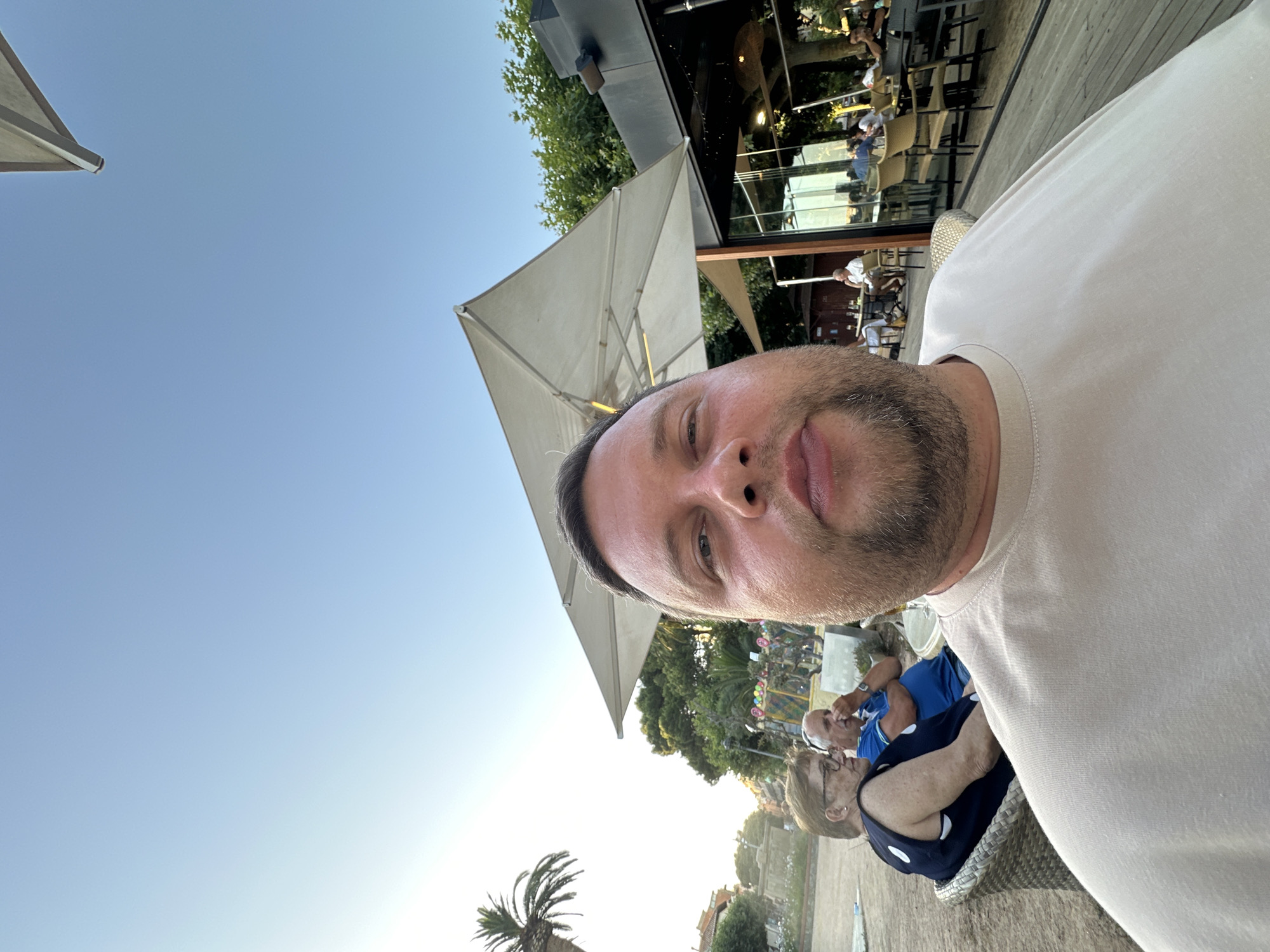 A man, positioned very close to the camera, takes a selfie at an outdoor seating area in Sant Feliu De Guíxols, Spain. He has a beard and is wearing a tan t-shirt. In the background, to the left, two individuals, an older woman with glasses and a man in a blue shirt, are seated at tables. The woman is wearing a dark sleeveless top with white polka dots. The man is looking away from the camera. Further in the background, trees and a building with large glass windows are visible. There are umbrellas providing shade over some of the seating areas. The sky is clear and blue.