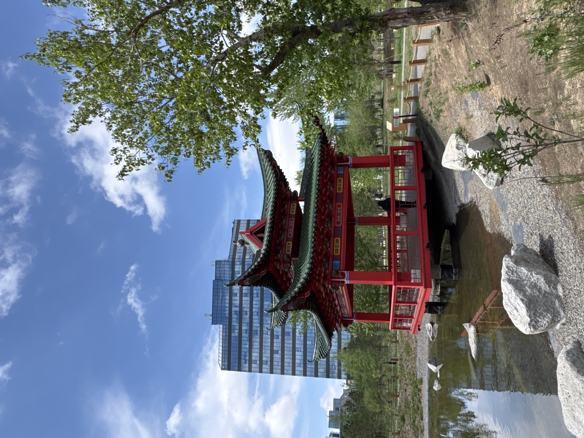 The image captures a serene scene in Ulaanbaatar, Mongolia, showcasing a traditional red and green Chinese-style pagoda situated next to a body of water. A modern, high-rise building is visible in the background, creating a juxtaposition of traditional and modern architecture. The pagoda is reflected in the water, adding depth and symmetry to the composition. The surrounding environment includes greenery, trees, and rocks along the water's edge, suggesting a park or garden setting. The sky is blue with scattered white clouds, indicating a clear day. The image appears to be taken from a low angle, emphasizing the height of the pagoda and the building behind it. The lighting is bright, casting reflections on the water and highlighting the colors of the pagoda.