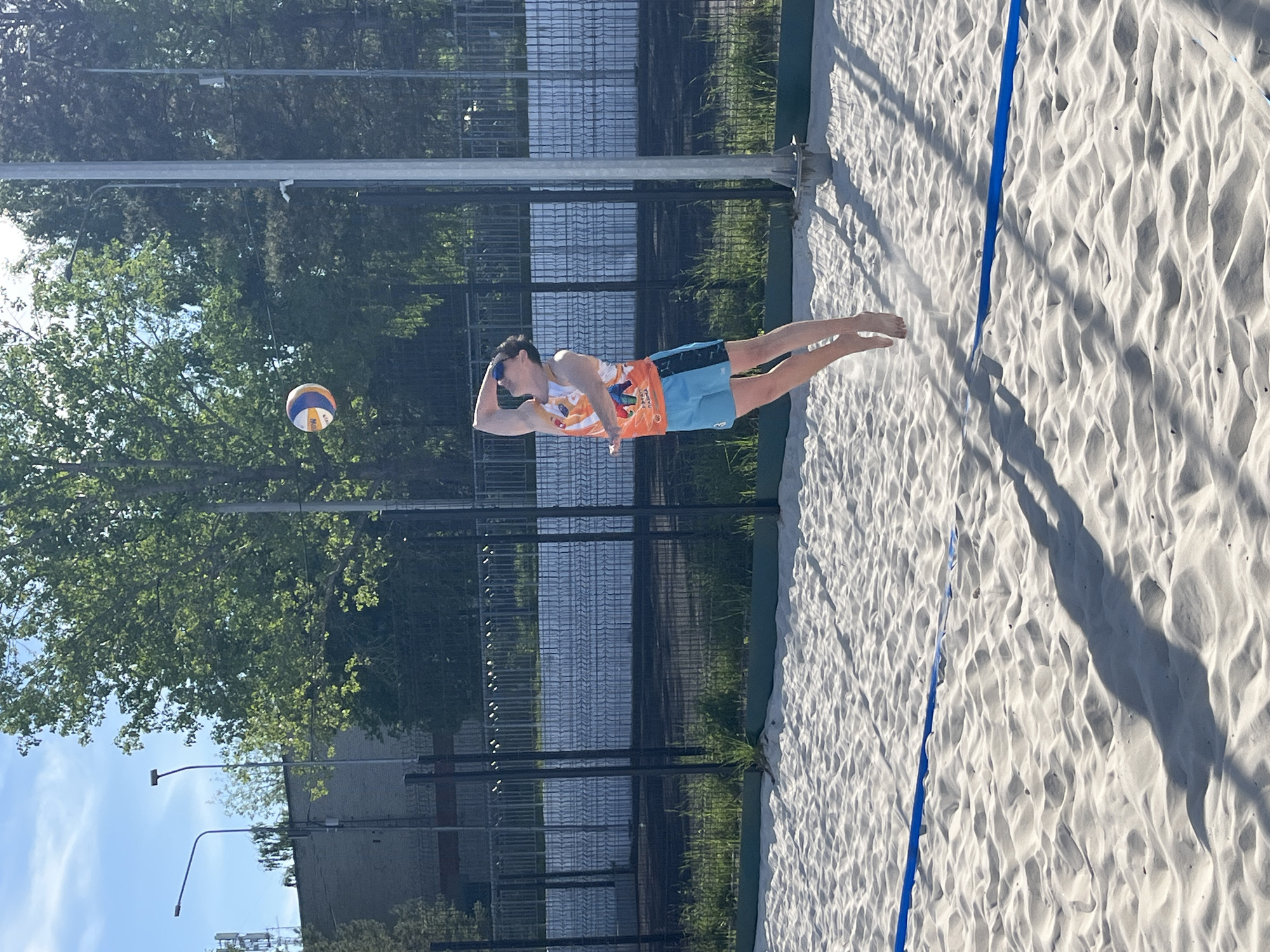A man dives for a volleyball on a sand volleyball court in Chaikovsky, Russia. The man is wearing sunglasses, a colorful patterned tank top, and blue and orange shorts. He is extended horizontally in mid-air, with his left arm bent at the elbow, presumably to make contact with the volleyball that is visible to the left of his head. The court is marked by a blue line running parallel to the man's body. A tall fence is visible in the background, beyond which are trees and a body of water. Lampposts are visible in the background.
