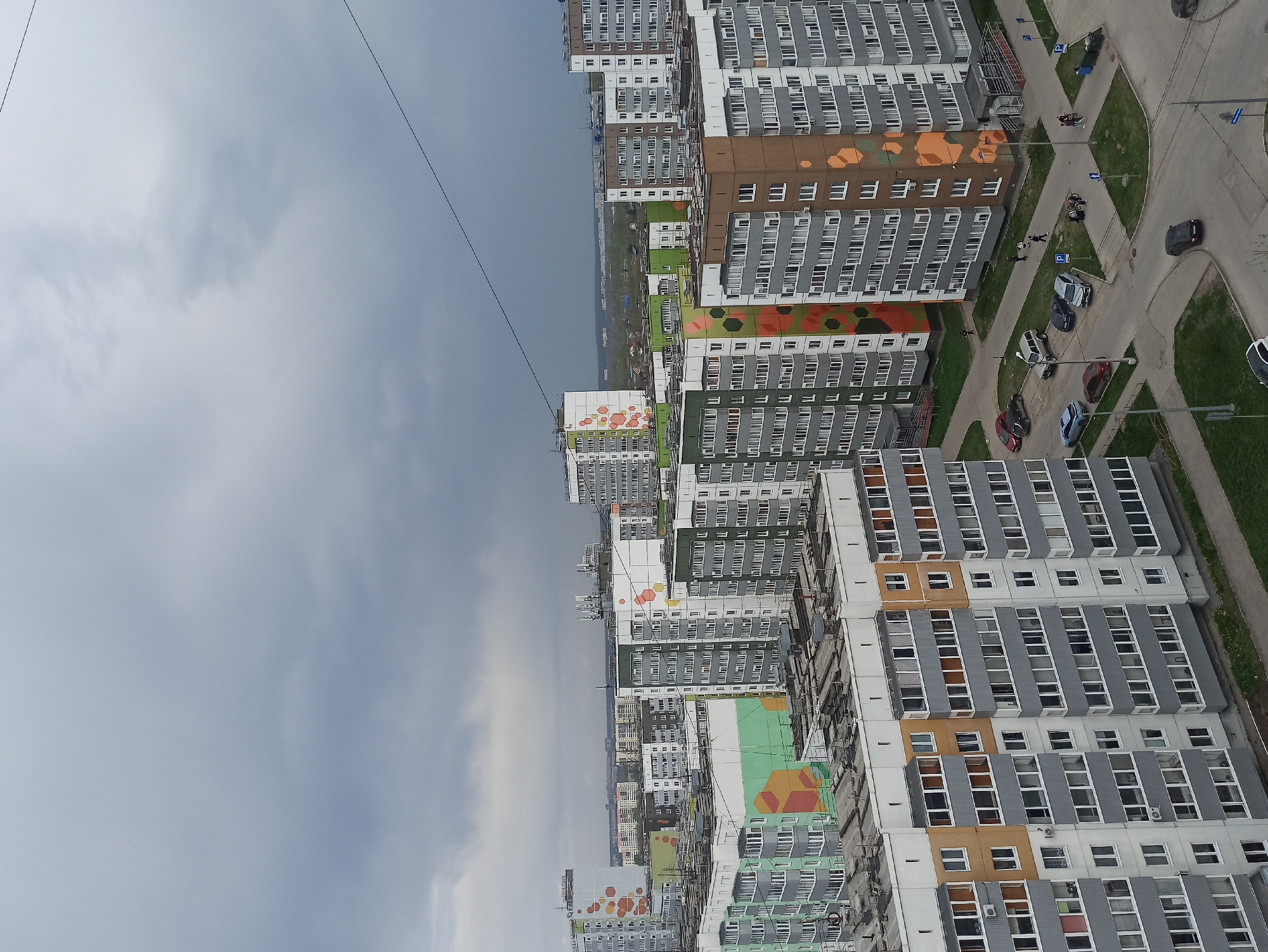 The image shows an aerial view of residential buildings in Кондратово, Russia, under a cloudy sky. The buildings are high-rise apartment complexes with multiple floors and numerous windows. Many of the buildings have colorful geometric patterns painted on their facades. Roads and sidewalks surround the buildings, and several cars are parked along the streets. Some pedestrians are visible on the sidewalks, interacting with the environment. A grassy area is adjacent to the road. A power line is visible in the sky.