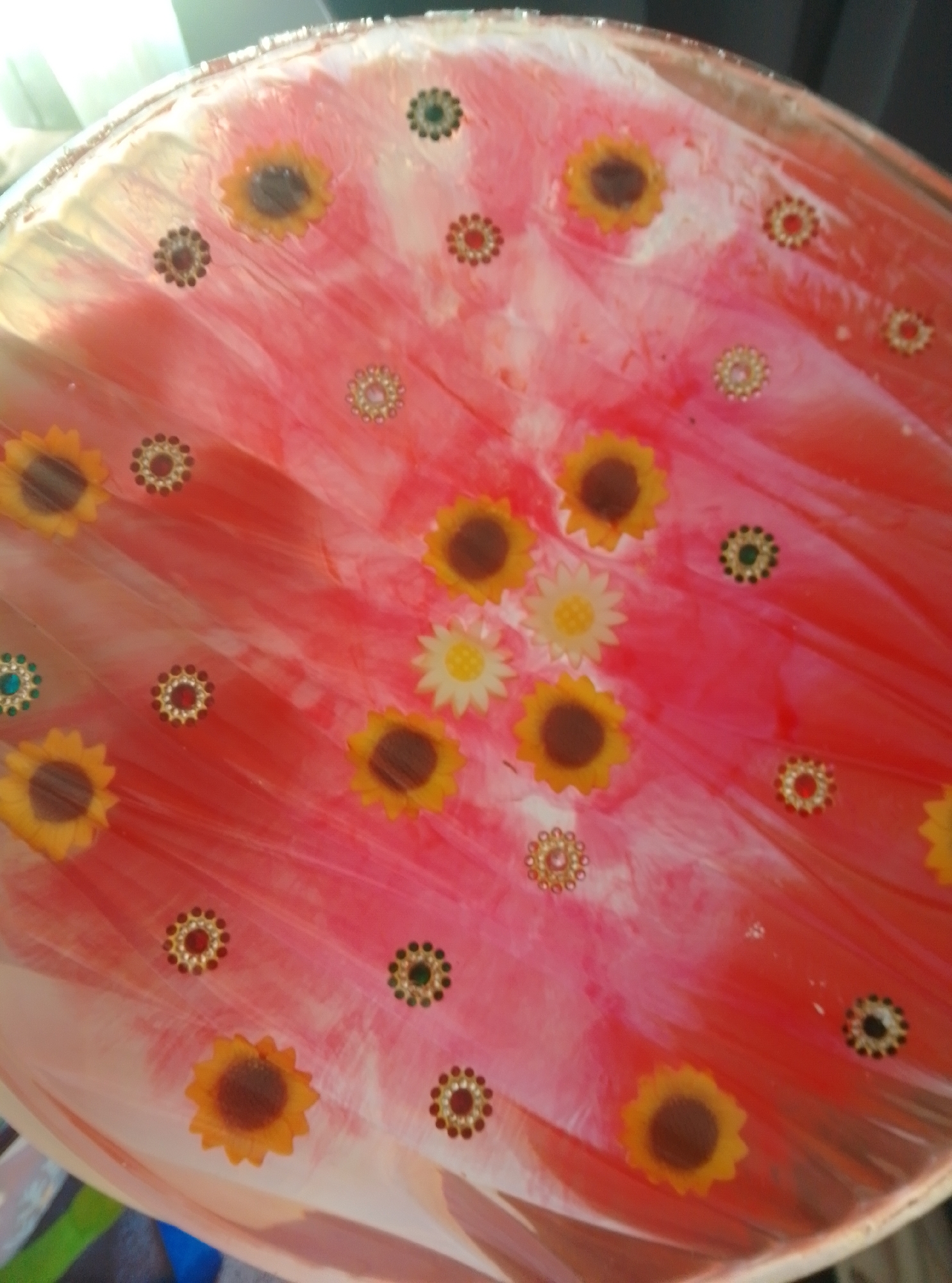 The image shows a close-up of a circular, resin-coated object with a pink and white swirled pattern. Scattered across the surface are numerous decorative elements resembling small flowers and floral designs. These decorations include yellow sunflowers with brown centers, white daisies with yellow centers, and circular embellishments with small red, green, or brown beads. The object has a slightly translucent quality, and the surface appears smooth and glossy. The lighting suggests the object is indoors, possibly under ambient light or diffused sunlight. There is no discernible location in the city of Wedel, Germany, visible in this image, as it focuses solely on the decorative surface of the object. No people, actions, or interactions are depicted.