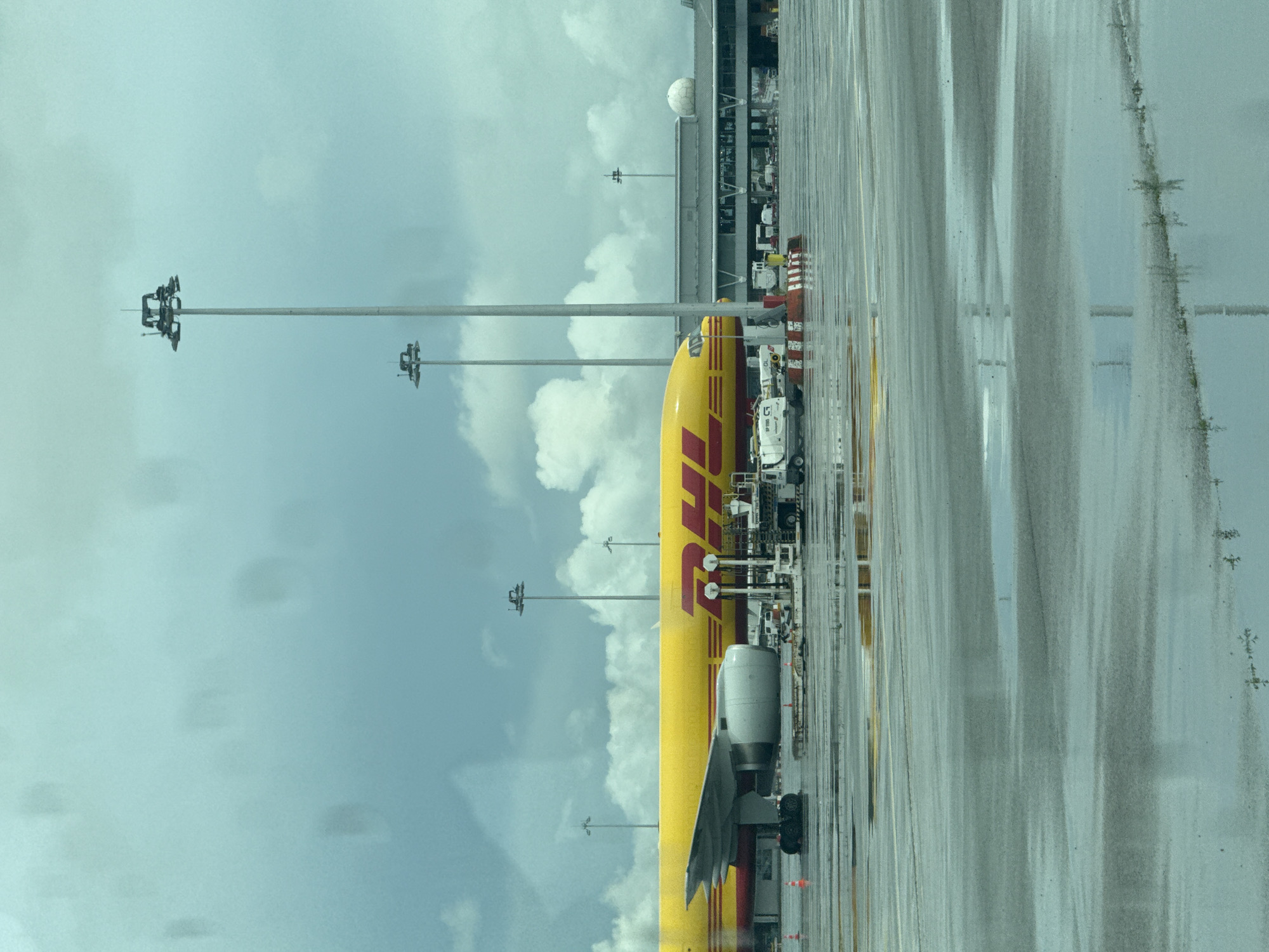 The image displays a DHL cargo airplane parked on a wet airport tarmac under an overcast sky during daytime. The aircraft, primarily yellow with a prominent red "DHL" logo on its fuselage, is seen from a slightly elevated vantage point. Its left wing, engine, and main landing gear are visible.
Several ground support vehicles, including what appears to be a fuel truck (with "SP 7000" visible on its side) and other equipment, are positioned near the plane's front section. Orange traffic cones mark areas on the tarmac. Multiple tall, high-mast floodlights are visible against the cloudy sky.
In the background, an airport terminal building with large windows and a white, spherical radome on its roof is present. The wet tarmac, reflecting the sky and surrounding structures, indicates recent rainfall. The sky is filled with light and dark grey clouds. The scene captures a moment of airport operations, likely in Sofia, Bulgaria.