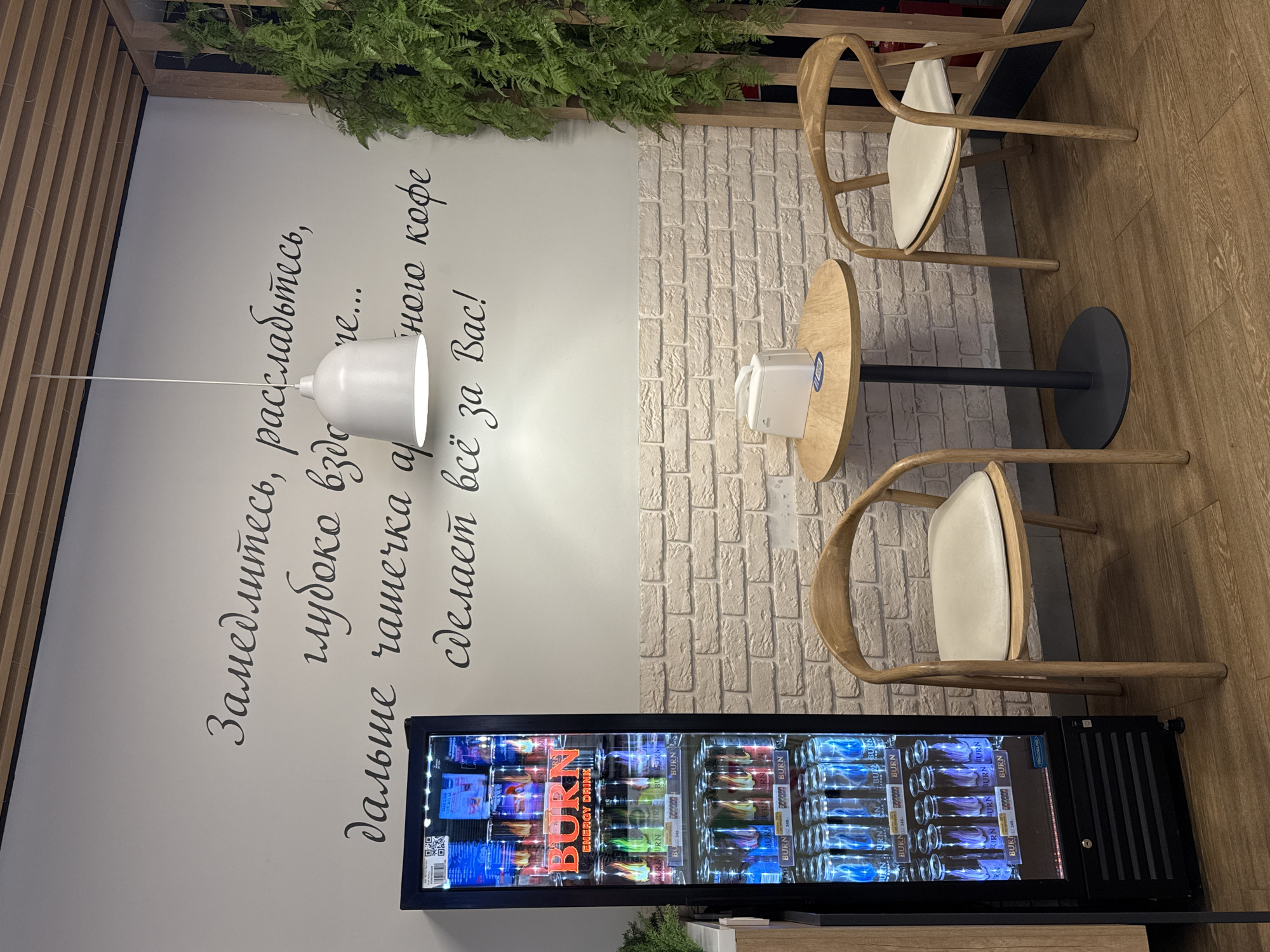 The image presents a cafe interior, possibly in Saint Petersburg, Russia, suggested by the Cyrillic text on the wall. The layout includes two light-colored wooden chairs positioned around a small, round wooden table with a black base. The table is currently unoccupied.
Adjacent to the table and chairs, on the left side of the frame, there's a wall featuring a decorative fern at the top and an inscription in Russian script. Below the inscription, there is a refrigerated display case filled with "BURN" energy drinks. The bottles are neatly stacked and illuminated, with the brand name clearly visible on the packaging.
The environment suggests a contemporary cafe setting, with a mix of materials such as wood, white brick-like paneling, and what appears to be a light-colored wood floor. The color palette is dominated by neutral tones, with pops of color from the energy drink cans. A modern white pendant lamp hangs above the setting.