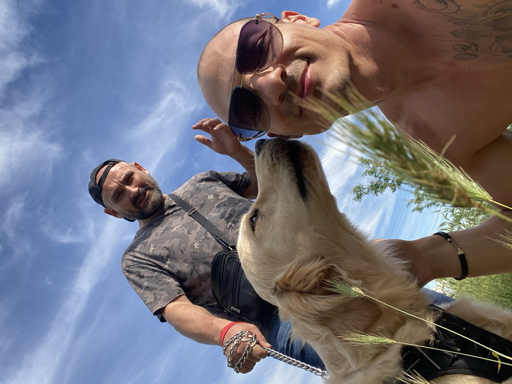 This is a close-up selfie taken outdoors in Pleven, Bulgaria. Two adult males and a golden retriever dog are present. One man, in the foreground and shirtless, wears aviator sunglasses and has tattoos on his chest. He is looking towards the camera, and the dog is sniffing his face. The second man, further back and wearing a camouflage t-shirt and a black baseball cap, is holding a chain leash attached to the dog. He is looking down and gesturing with his left hand. The background shows a blue sky with white clouds and some green foliage, suggesting a park or natural area.