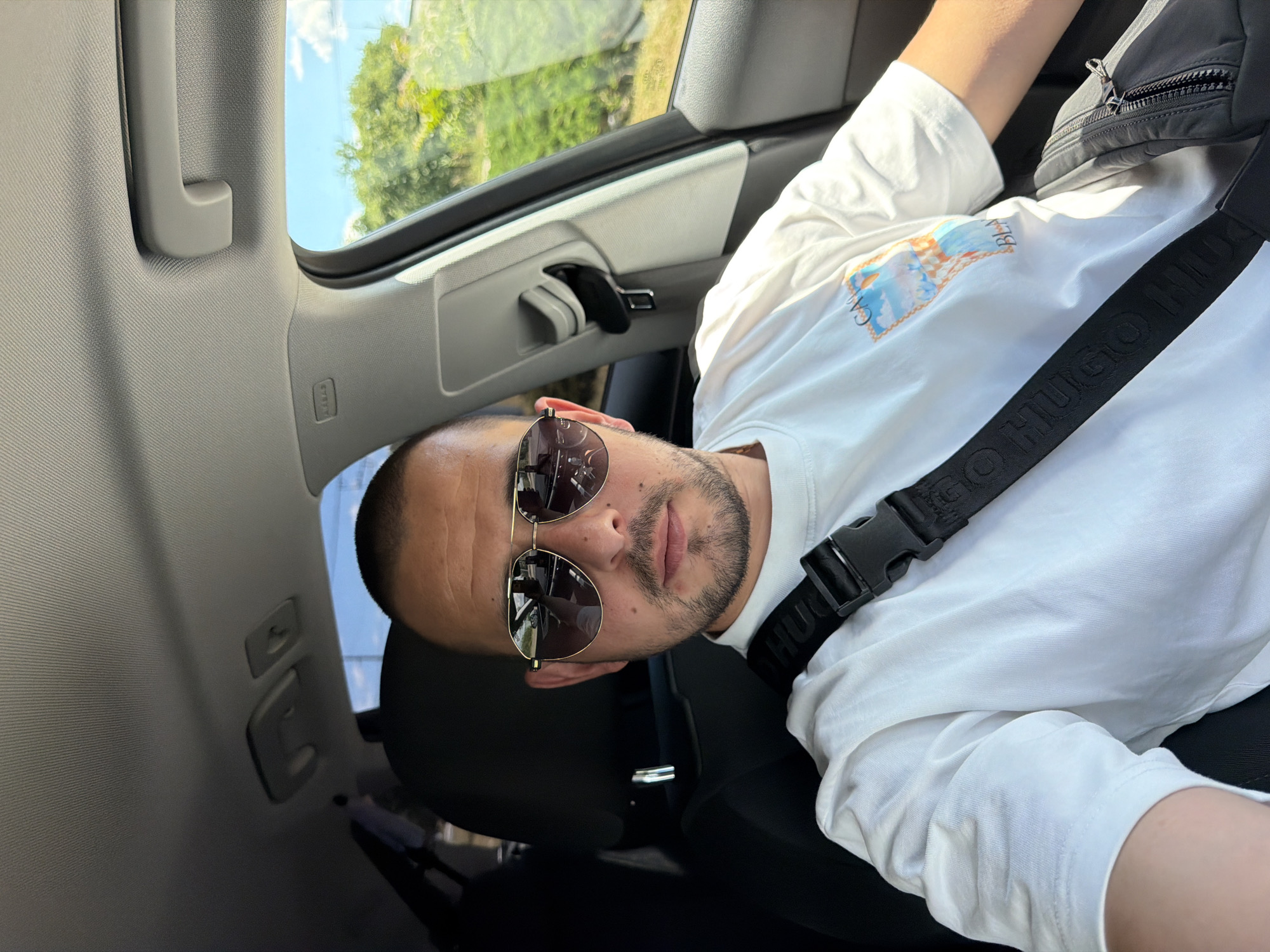 A male with short brown hair and stubble is sitting in the driver's seat of a car, wearing aviator sunglasses and a white t-shirt with a graphic print. A black seatbelt crosses his chest, and a black bag is visible on his right side. The interior of the car, including the dashboard, steering wheel, and a grab handle, is visible. Outside the car window, lush green trees and a blue sky with clouds are seen, suggesting a sunny day. The image appears to be taken in Burgas, Bulgaria, judging by the visible landscape.