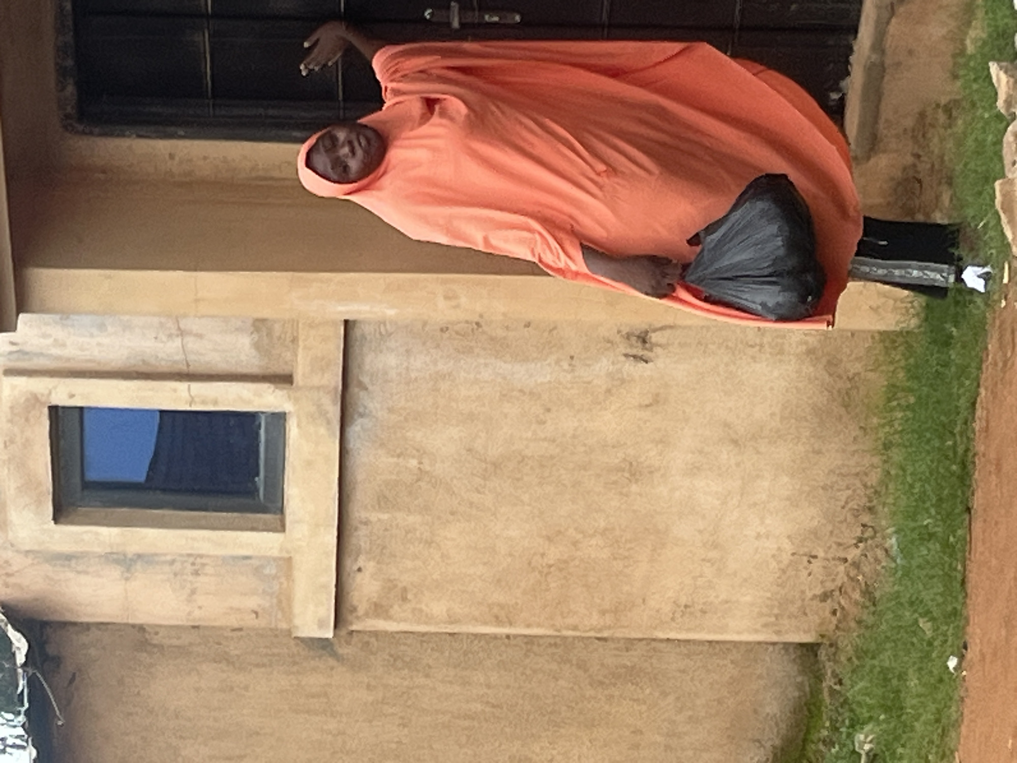 A medium shot shows a young Black woman standing outside a building with tan stucco walls and a dark door. She is wearing a flowing coral-colored hijab and dress, and black pants. She is holding a black plastic bag in her right hand. Her left hand is raised, with her palm facing forward, and she is looking toward the camera. The building has a small, dark-paned window above and to the left of the woman. In the foreground, green grass with some litter is visible, as is a dirt path. The overall scene suggests a residential area in Bukuru, Nigeria.