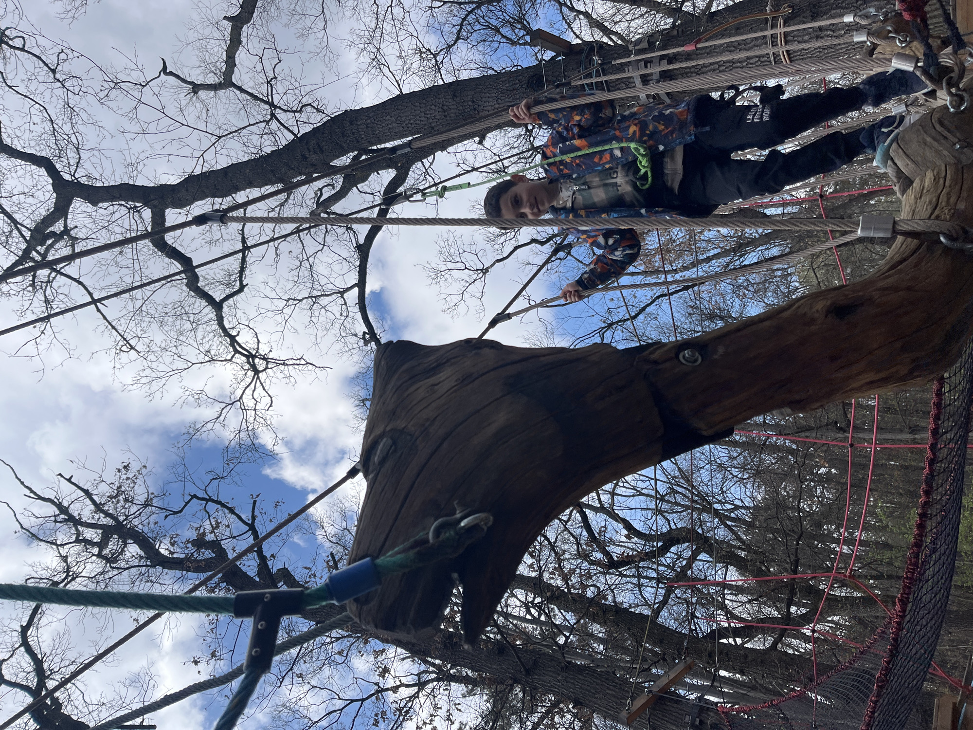 A low-angle shot depicts a child navigating a high ropes course, likely located outdoors. The child, wearing a patterned jacket and dark pants, is positioned on a rope bridge, holding onto the ropes for support. Below, a large wooden structure resembling a tree stump with attached ropes and hardware is visible. Overhead, bare tree branches reach towards a sky with scattered clouds. Although this information is not discernable from the image, the location is stated to be in Sofia, Bulgaria. The focus of the scene is on the child and the elements of the ropes course.