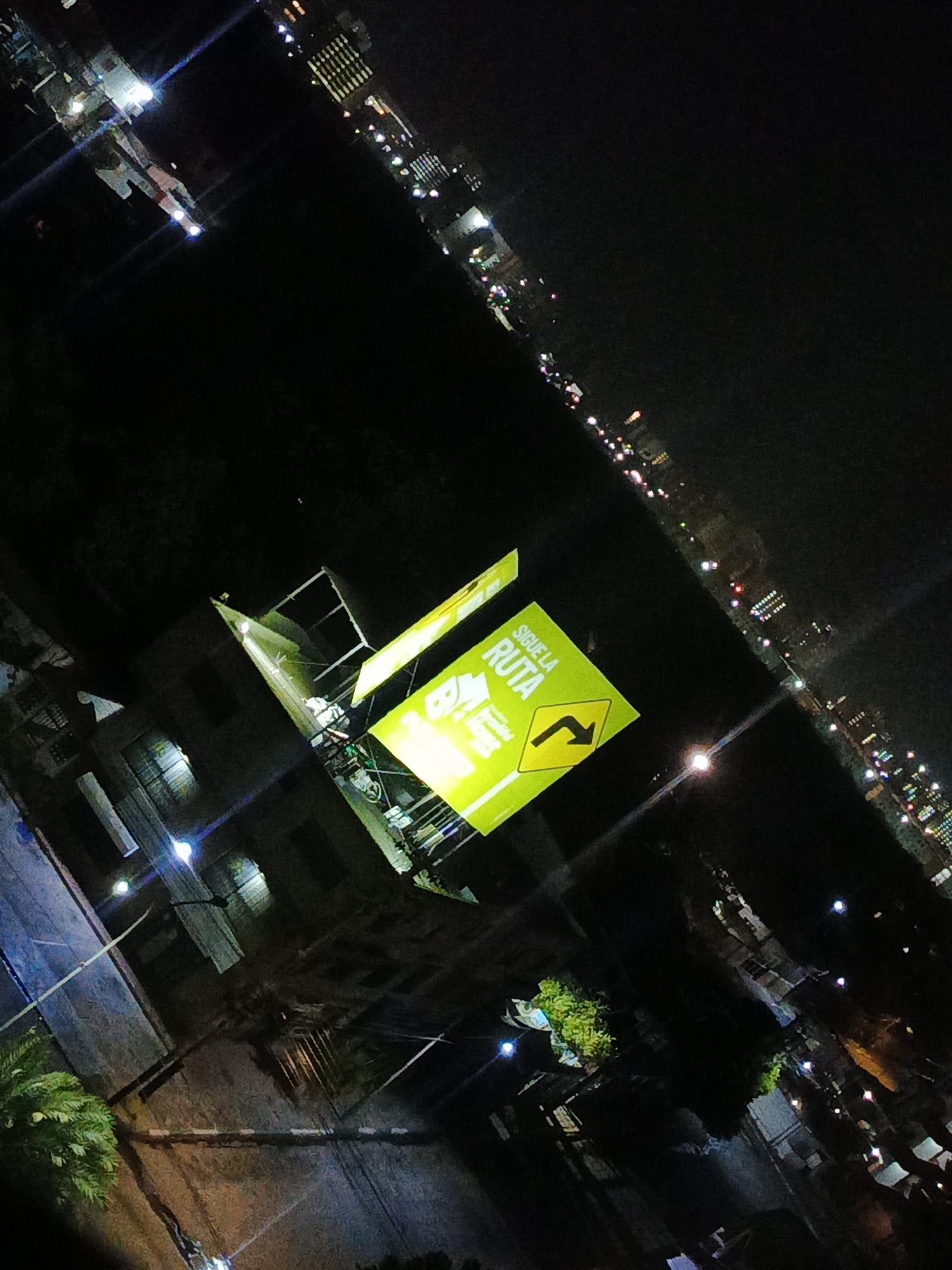 At night, the city of Los Robles, Venezuela, is dimly lit. In the foreground, a brightly lit billboard displays a yellow sign with the words "SIGUE LA RUTA" and a right-turning arrow. The billboard is attached to a building and illuminated by streetlights. The surrounding environment is dark, with sparse vegetation and scattered streetlights. In the distance, a line of buildings with lights outlines the city's horizon.
