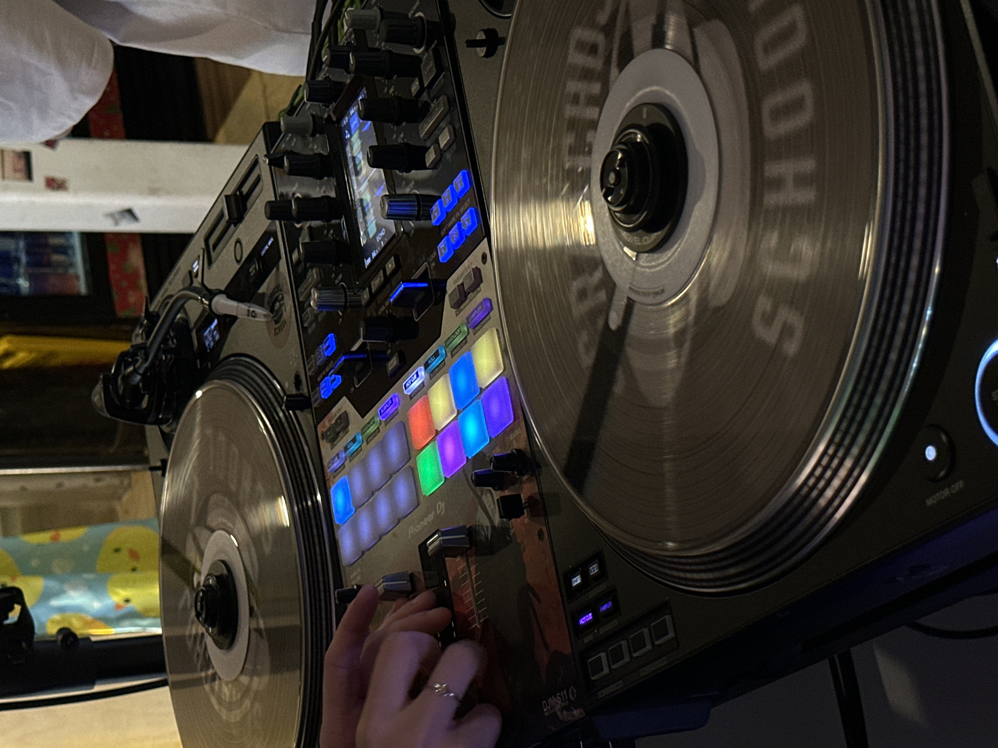 The image displays a close-up, high-angle shot of a DJ's setup. In the foreground, a turntable with a record is visible, the record displaying the text "SCHOOL" in a stylized font. To the left, a person's hands are manipulating controls on a DJ mixer. The mixer features numerous buttons, sliders, and illuminated pads in various colors, including blue, green, orange, and yellow. The brand name "Pioneer DJ" is discernible on the mixer. In the background, another turntable is present, and a portion of a person wearing a white t-shirt is visible on the right. The background is dimly lit and shows a blurred shelf with bottles. The overall scene suggests an indoor setting, possibly a club or event space. There is no specific information in the image to place it in Khimki, Russia.