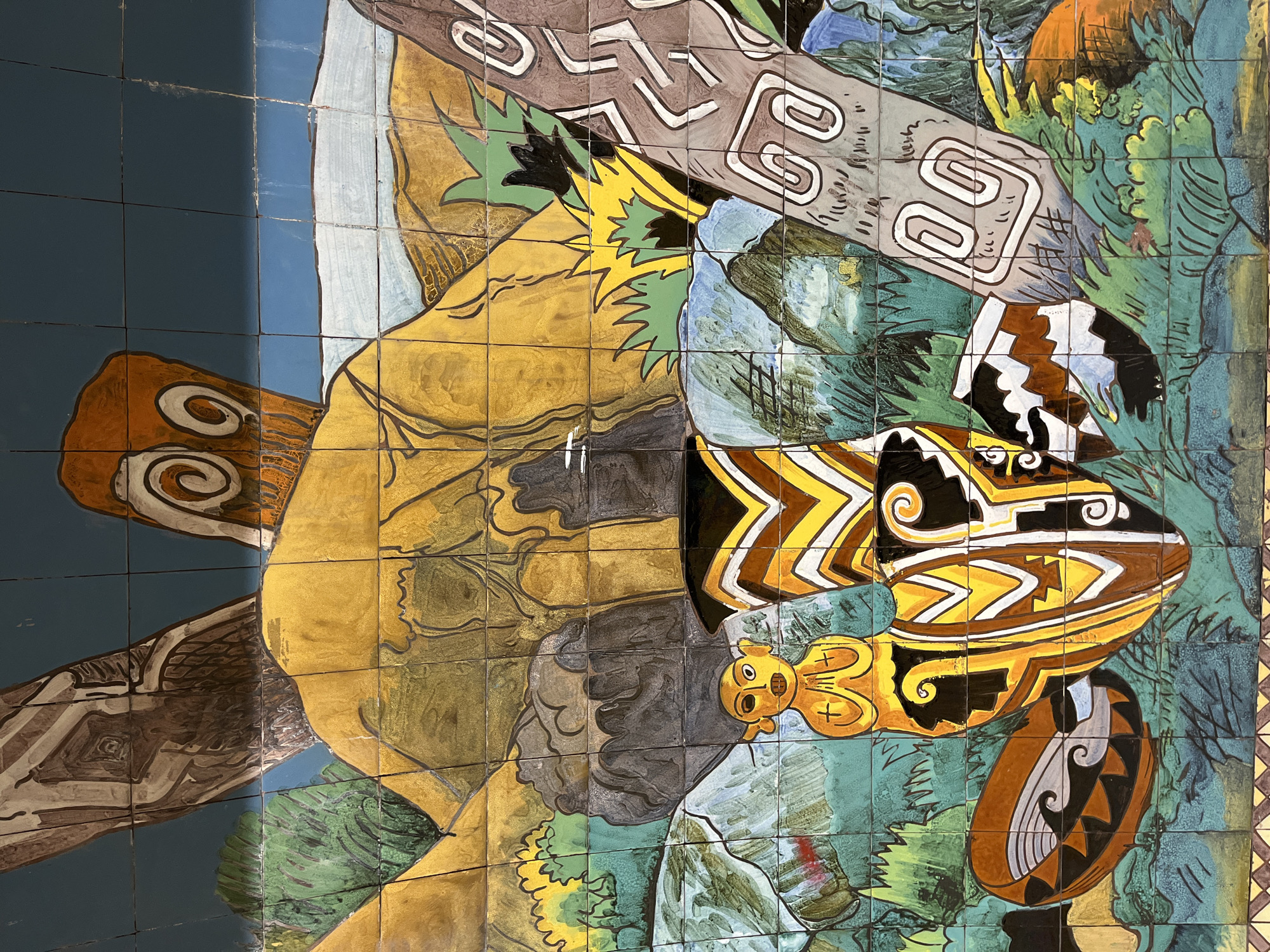 This media file captures a close-up of a vibrant, multi-colored mural composed of numerous ceramic tiles, likely a public art installation in Buenos Aires, Argentina. The artwork features highly stylized, possibly mythological or indigenous figures and natural elements.
A large, dominant figure, rendered primarily in shimmering gold and shades of brown and yellow, occupies the central and left-hand portions of the composition. Its form is organic and somewhat anthropomorphic, with defined lines and contours. Below and to the right of this golden figure, a smaller, bright yellow figure with distinct facial features (eyes, nose, mouth) and cross-like markings on its body is visible.
Surrounding these central figures are elements suggestive of a natural environment. Dark and light blue sections likely represent water, while various shades of green and teal depict dense foliage or landscapes. Several large, elongated shapes resembling logs or tree trunks, decorated with white spiral patterns, are integrated into the design. Other distinct objects, possibly stylized fish or canoes, are painted with intricate black, white, brown, and yellow zigzag and spiral motifs.
The overall aesthetic points to South American indigenous art traditions, characterized by bold lines, rich colors, and symbolic imagery. The individual ceramic tiles contribute a textured, segmented appearance to the mural. There is no visible text within the image, and the scene depicts a static narrative or symbolic representation rather than an active event.