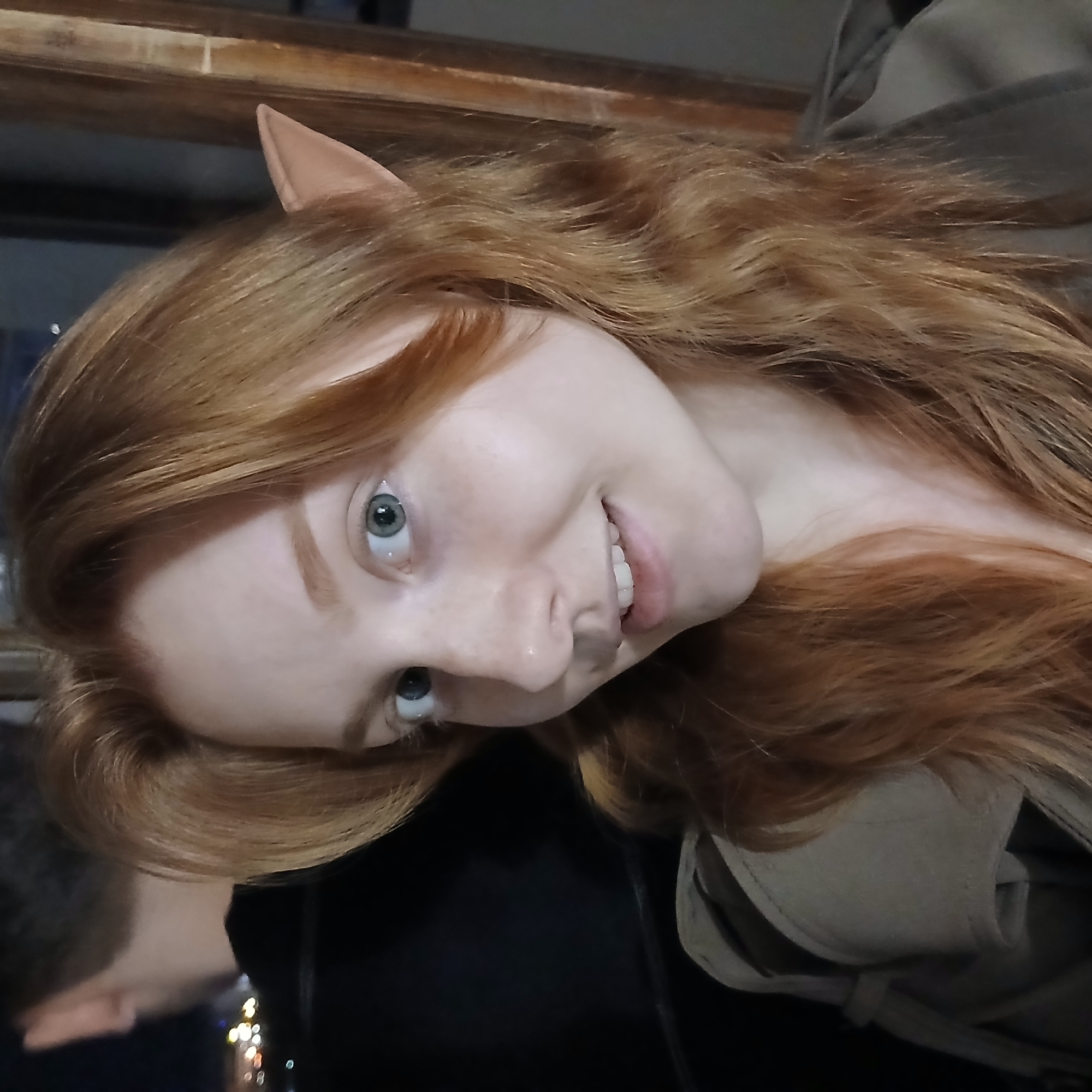 A close-up, tilted portrait captures a young woman with fair skin, prominent light blue-green eyes, and long, wavy reddish-blonde hair. She wears a light brown, pointed elf ear prosthetic on her right ear, suggesting a costume or fantasy theme. A slight smile reveals her teeth as she looks directly at the camera. She is dressed in a light olive green or beige coat or jacket.
The background features a dark wooden beam horizontally across the top, beneath which is a dark, reflective surface. In the lower background, blurred, colorful lights are visible, indicating an evening or nighttime setting, possibly indoors or overlooking a city. A partial reflection in the bottom left shows the back of another person's head with darker hair. The woman's expression is pleasant and engaging. The image was taken in Yekaterinburg, Russia. No visible text is present.