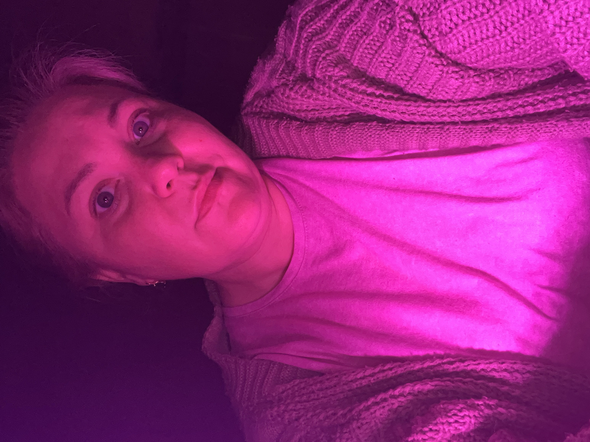A woman is shown in a selfie-style image with pink lighting. She is wearing a pink t-shirt and a chunky knit cardigan. Her expression is somewhat pensive. The location in the city of Vesyoloye, Russia, cannot be determined from the image.