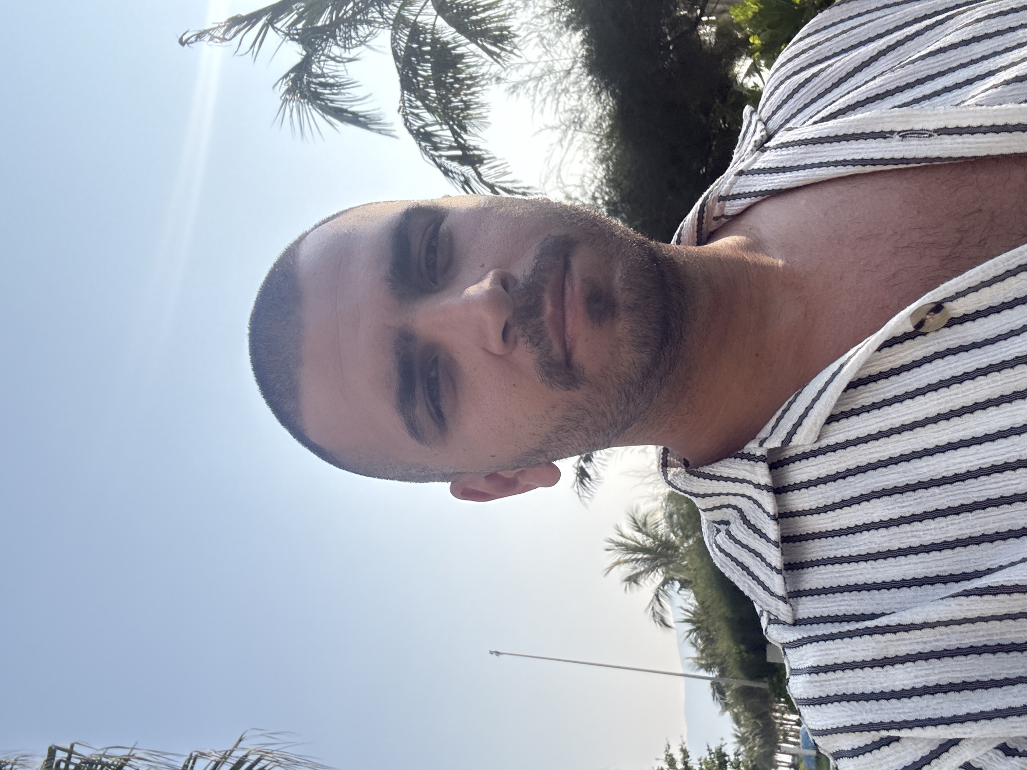The image is a selfie of a man with short, dark hair and a beard, wearing a striped shirt. The background shows a bright, sunny day with palm trees and a clear blue sky. Based on the environment, the location is likely Đà Nẵng, Vietnam. There is a pole in the distance, suggesting a street or coastline setting. The man is facing the camera, and the selfie is taken from a low angle.