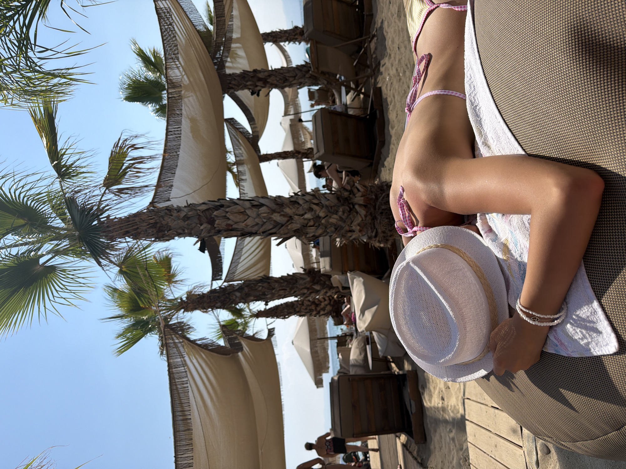 In Ofrynio Beach, Greece, a person lies on a lounge chair on a sandy beach. They are wearing a white hat and a pink and white checkered bikini. Their back is to the camera, and their head is turned away, obscured by the hat. Several palm trees and beach umbrellas are visible in the background, suggesting a sunny day at a resort. The person appears to be relaxing or sunbathing.