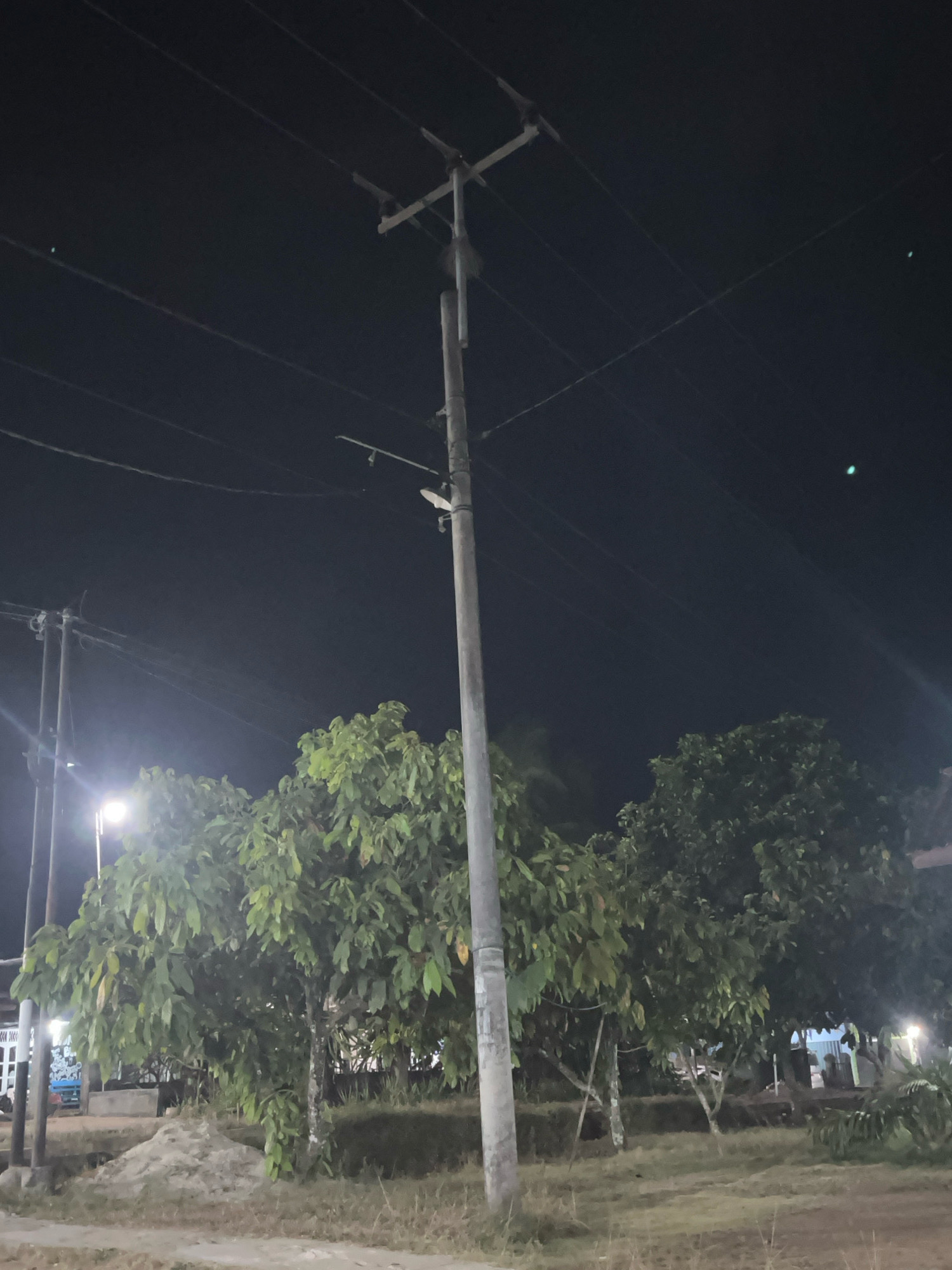 This is a nighttime photograph taken in Kebumen, Indonesia. The image features a prominent utility pole with multiple wires stretching across the dark sky. In the foreground, lush green trees with large leaves are visible, partially illuminated by distant lights. The ground is covered with dry grass and some loose soil. In the background, there are hints of structures and streetlights, suggesting an outdoor, possibly public, area. The overall scene is dimly lit, typical of a rural or suburban setting at night.