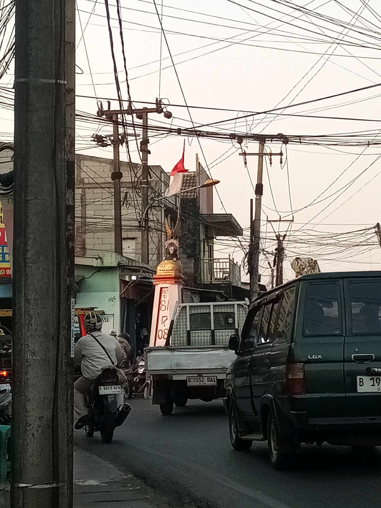 The image depicts a street scene in Karangsatria, Indonesia, characterized by a dense network of overhead electrical wires. In the foreground, a motorcycle with a rider wearing a helmet and a light-colored shirt is moving away from the camera. Behind the motorcycle, a white truck with a license plate reading "B 9552 OAI" is visible. To the right, the rear of a dark green SUV with the "LGX" logo is in the lane. Further in the background, a monument or marker with a red and white flag on top stands near buildings and more utility poles. The sky is overcast, suggesting it is either early morning or late afternoon. The overall environment appears to be a typical urban or semi-urban street with significant traffic and infrastructure.