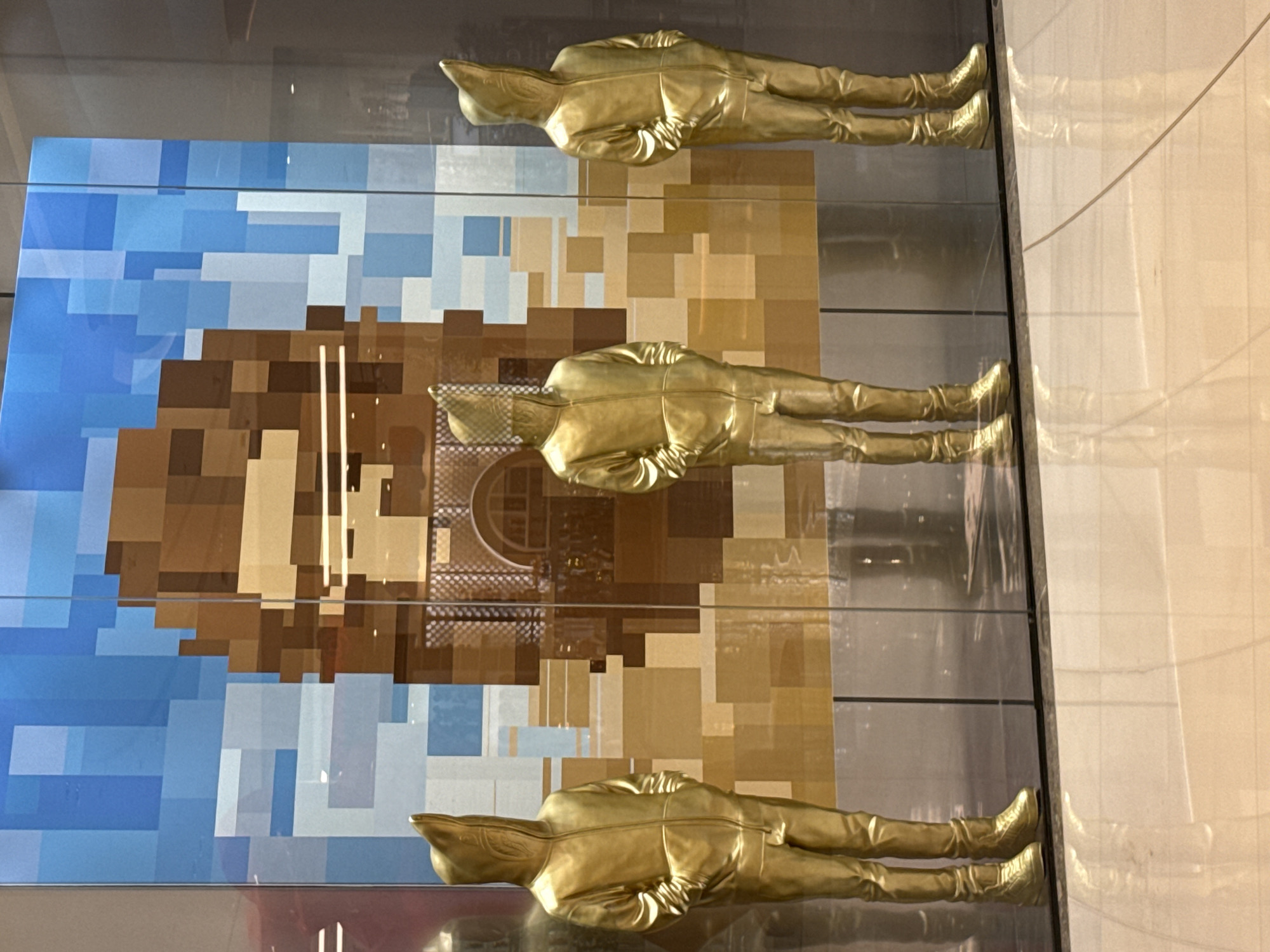 Three gold-colored mannequins dressed in hooded zip-up jackets and pants stand in front of a large, pixelated mural in a glass-enclosed display. The mural features a brown and tan abstract design with blue hues at the top, possibly depicting a sky or landscape. The mannequins are positioned symmetrically, with their hands in their pockets. The foreground shows a reflective tiled floor. The scene appears to be a storefront or exhibition space, likely in a public area or mall. The style of the mannequins and mural suggests a contemporary art or fashion display. The location is in Dubai, United Arab Emirates.