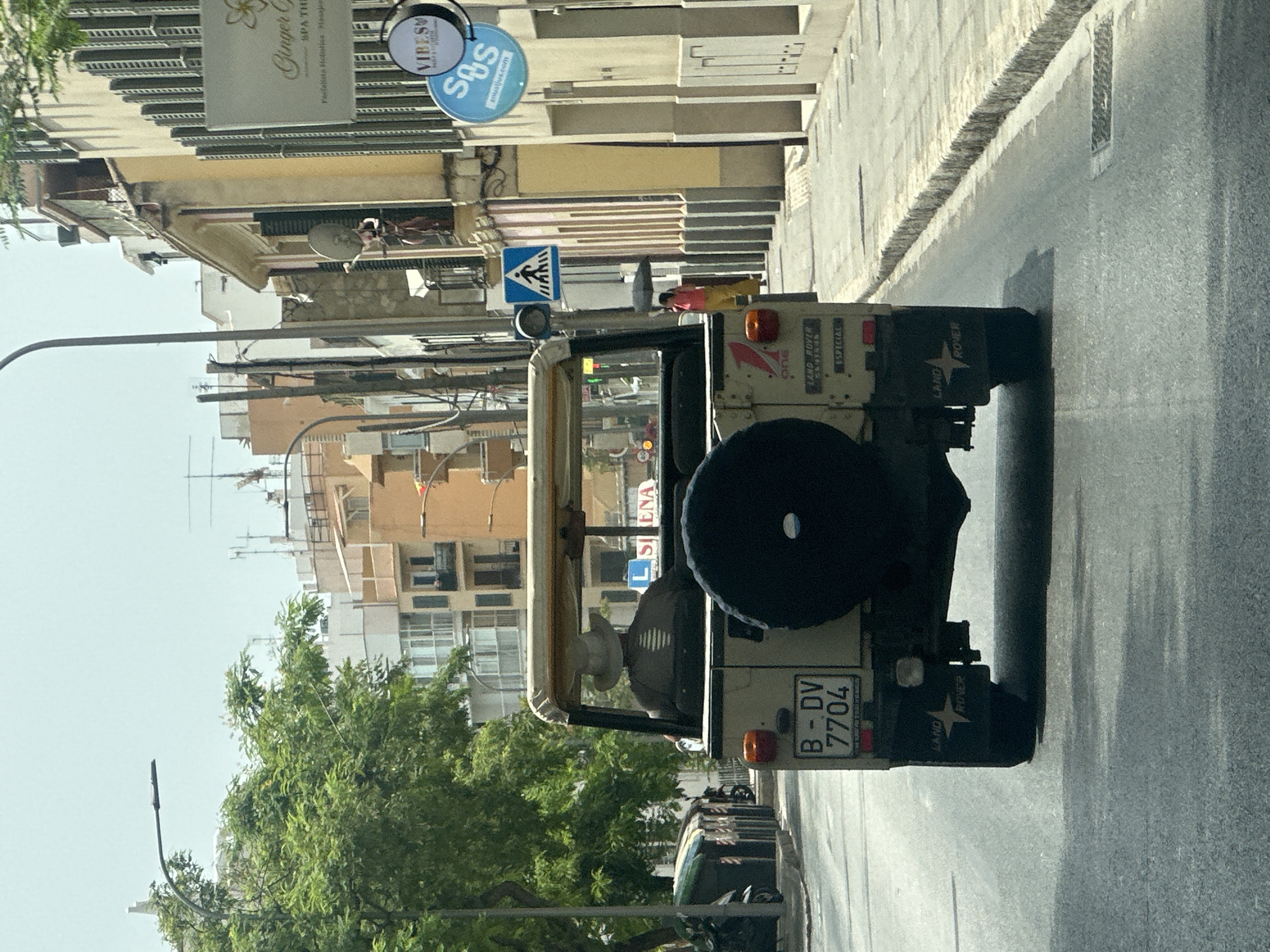 The image shows a view from behind a beige-colored open-top Land Rover vehicle driving on a street in Palma de Mallorca, Spain. The vehicle has a spare tire mounted on the back and a license plate that reads "B-DV 7704". The driver, wearing a light-colored hat, is visible in the driver's seat. The street is paved and lined with buildings on the right side, which appear to be shops or residences. One building has a sign that reads "Ginger C" and another sign with "SOS". A pedestrian crossing sign is visible on a pole. A person holding a black umbrella is walking on the sidewalk to the right of the vehicle. Trees and more buildings are visible in the background, under a bright, overcast sky. The overall scene depicts a typical urban streetscape in a Mediterranean city.