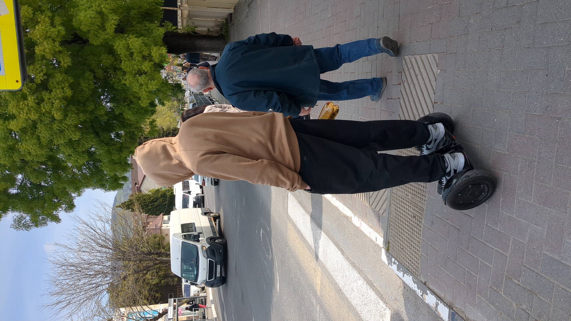 The image captures a scene in what appears to be the city of Gelendzhik, Russia, as identified by the license plate on a parked vehicle in the background. Two individuals are visible on a paved sidewalk. One person, wearing a tan hoodie and black pants, is riding a self-balancing scooter. Their feet are clad in grey and black sneakers. Beside them, an older man in a blue jacket and jeans walks alongside. Street markings are visible. In the background, vans are parked, and trees line the street. The sky is partly cloudy.