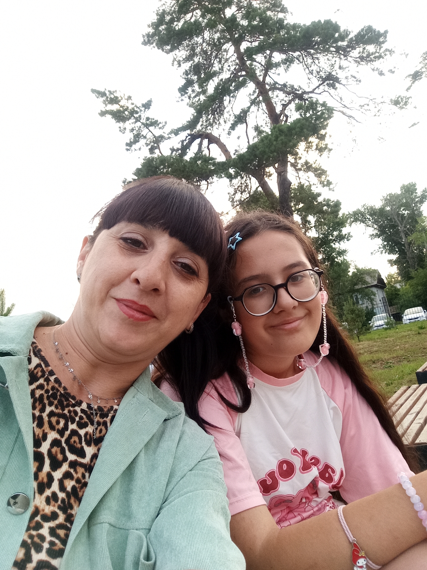 This media file is a selfie of two females, likely a mother and daughter, sitting outdoors on a sunny day. The older female, in the foreground on the left, has dark hair with bangs and is wearing a leopard print top and a light green jacket. She is smiling and looking at the camera. The younger female, to the right, has long brown hair and is wearing glasses with pink beaded chains attached. She is also smiling and looking at the camera. She has a light pink shirt with a graphic print and a bracelet on her wrist.
Behind them, a large tree with green foliage is visible against a bright sky, suggesting it's daytime with potentially overcast or hazy conditions. In the background, there are more trees, a building, a fence, and two cars parked on a grassy area. The overall setting appears to be a park or a rural outdoor space. The smiles on both individuals' faces suggest a happy and relaxed atmosphere. There is no discernible text in the image.