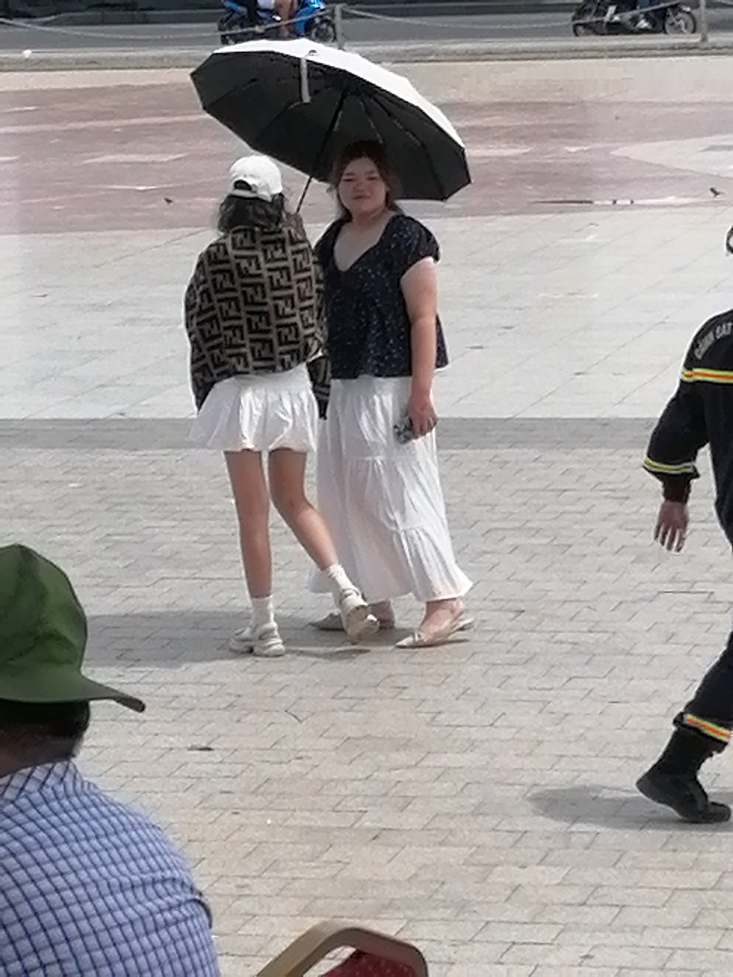 The image depicts an outdoor scene in Da Lat, Vietnam, likely on a paved pedestrian area with intersecting cobblestone-like patterns. Two women are the primary subjects. The woman on the left is wearing a white baseball cap, a patterned jacket with a repeated "F" logo, and a short white pleated skirt, paired with white socks and chunky white sneakers. The woman on the right is wearing a dark, short-sleeved top with a V-neck and a long white tiered skirt, also wearing light-colored flat shoes. They are standing together under a black umbrella. To the right, a person in a dark uniform with yellow and red stripes on the sleeves is visible from the chest down, wearing black boots. In the foreground, the back of a person wearing a blue and white checkered shirt and a green hat is visible, seated. The background shows a street with a motorcycle and other people. The overall impression is of a casual gathering or walk in a public space.