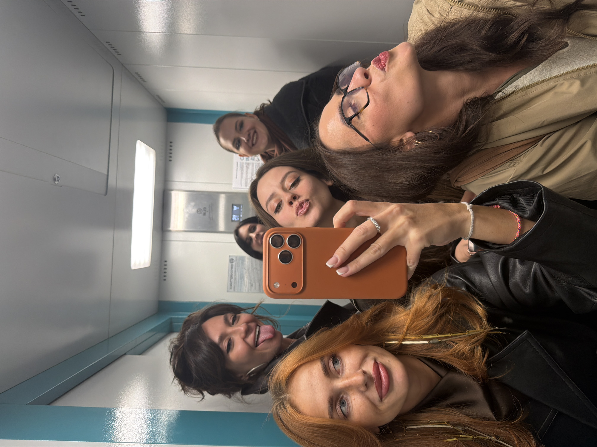 A selfie taken by one of six women in an elevator. The elevator is a pale gray and blue color with a light panel. A control panel is visible. The women are arranged closely, and all face the camera. From the bottom, clockwise, the woman at the bottom has red hair and wears a black leather jacket. The next woman has short dark hair and is sticking her tongue out. Above them, the woman holding the phone has long brown hair and is wearing a black leather jacket. The next woman has long brown hair and is kissing the camera. The next woman has dark hair and is smiling, and the last woman has brown hair and wears glasses, pursing her lips.
