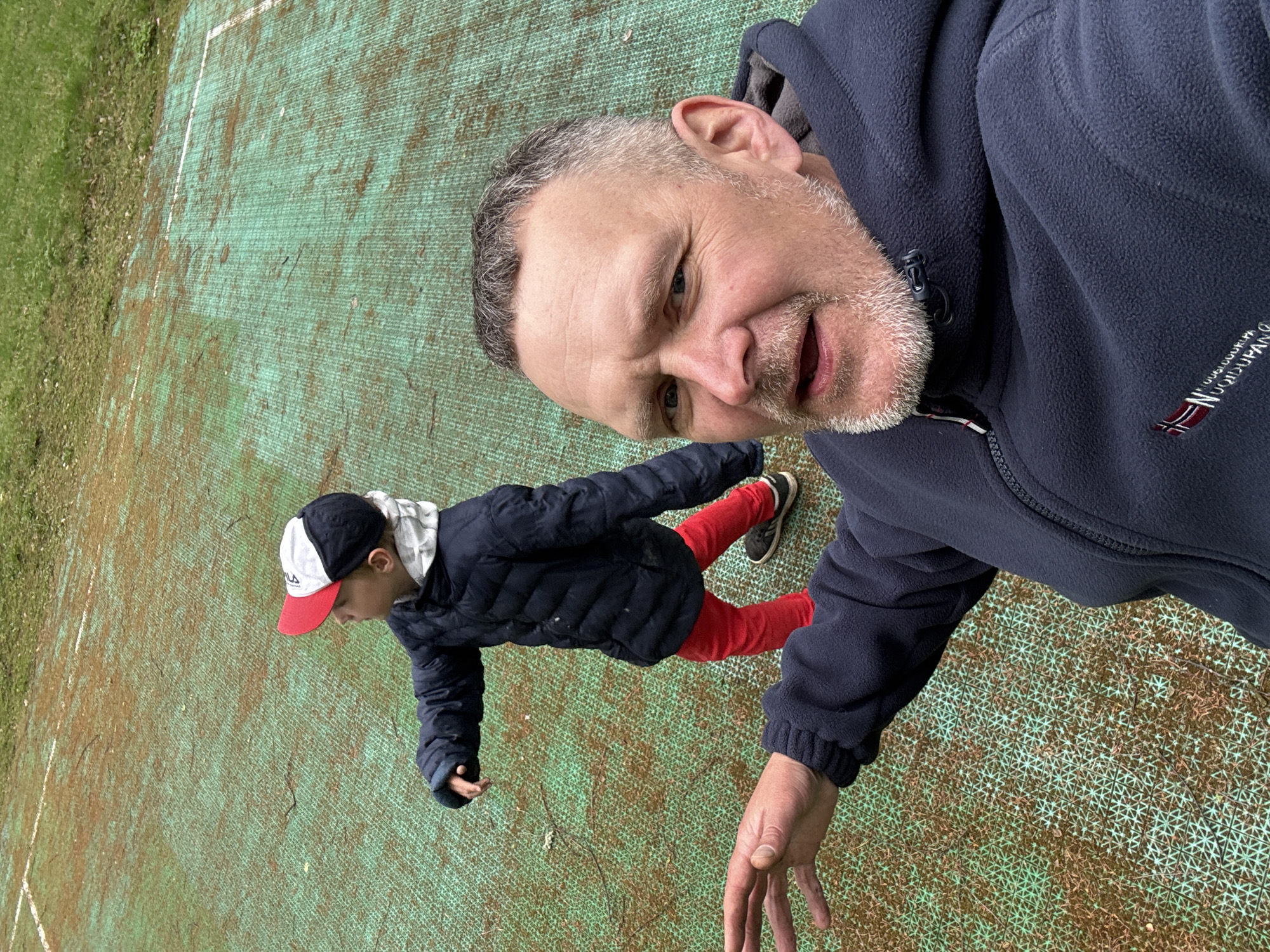 A man and a boy are photographed in Aleksandrovka, Russia. The man wears a navy blue fleece jacket and has graying hair and a beard. The boy wears a navy blue jacket, red pants, and a baseball cap with a white front and red brim. They stand on a green surface with white lines, possibly a sports court. The man appears to be taking a selfie, as his face is close to the camera and his hand is extended. The boy stands beside him.
