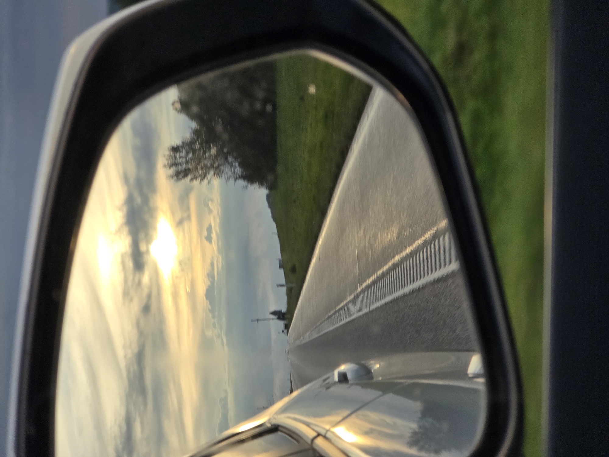 A side-view mirror of a car reflects a road stretching into the distance under a cloudy sky. The sun is visible in the upper left portion of the mirror, casting a warm glow on the clouds and the wet pavement of the road. Green grass lines the side of the road, and a few trees are visible in the background. The side of the car is also reflected in the mirror, showing its metallic surface. The image appears to be taken from inside the car while it is in motion. The scene takes place on a road in Prienai, Lithuania.
