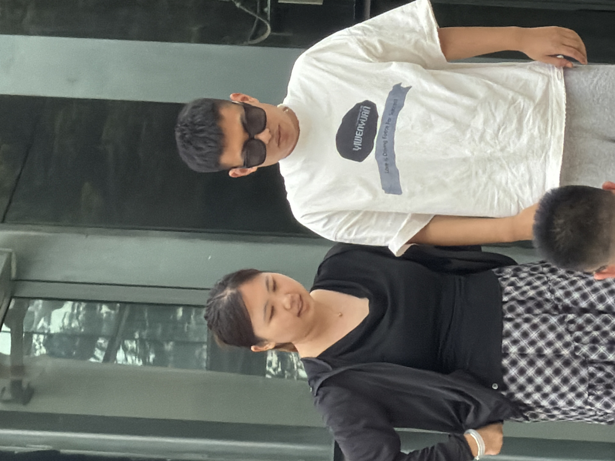 Two people, a woman and a man, are standing outdoors in front of a modern building with large glass windows. The woman, on the left, is wearing a black top and a black zip-up jacket, with a patterned skirt. She has dark hair tied back and is wearing a necklace. The man, on the right, is wearing a white t-shirt with a graphic print and sunglasses. He has short dark hair. The building's facade is reflective, showing glimpses of trees and the sky. The ground is paved. The scene appears to be in Yuzhong District, China, based on the provided context.