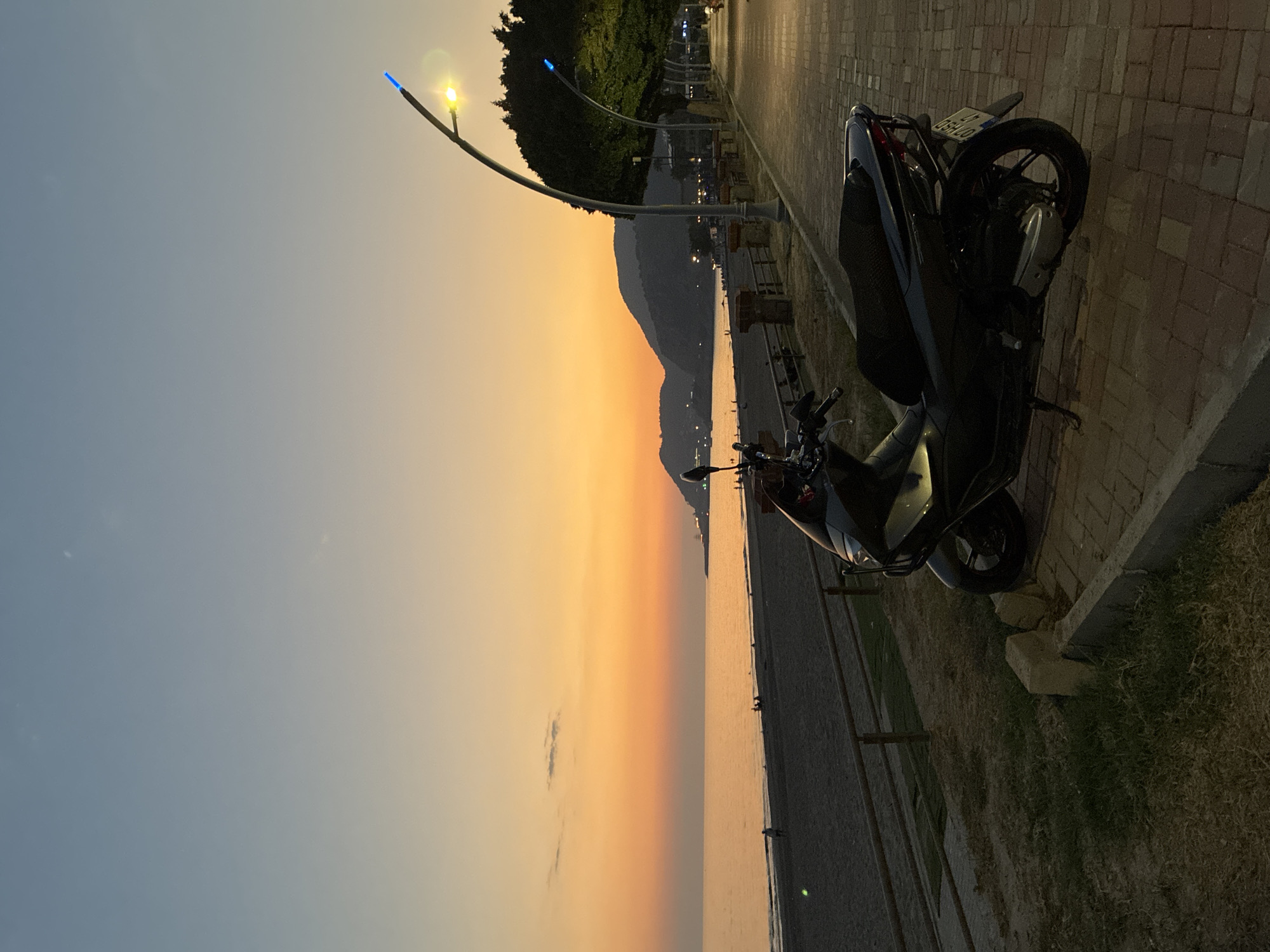 A dark-colored scooter is parked on a brick-paved walkway adjacent to a sandy beach and the sea, during sunset. The sky displays a gradient of orange and blue hues, with faint clouds. The sea is calm, reflecting the warm colors of the sky. Several people are visible on the beach and along the walkway. Modern streetlights with blue and yellow lights illuminate the pathway. In the background, hills or mountains are silhouetted against the twilight sky. The location is the coastal area of Alanya, Turkey, likely along the promenade. The scene captures a peaceful evening atmosphere.