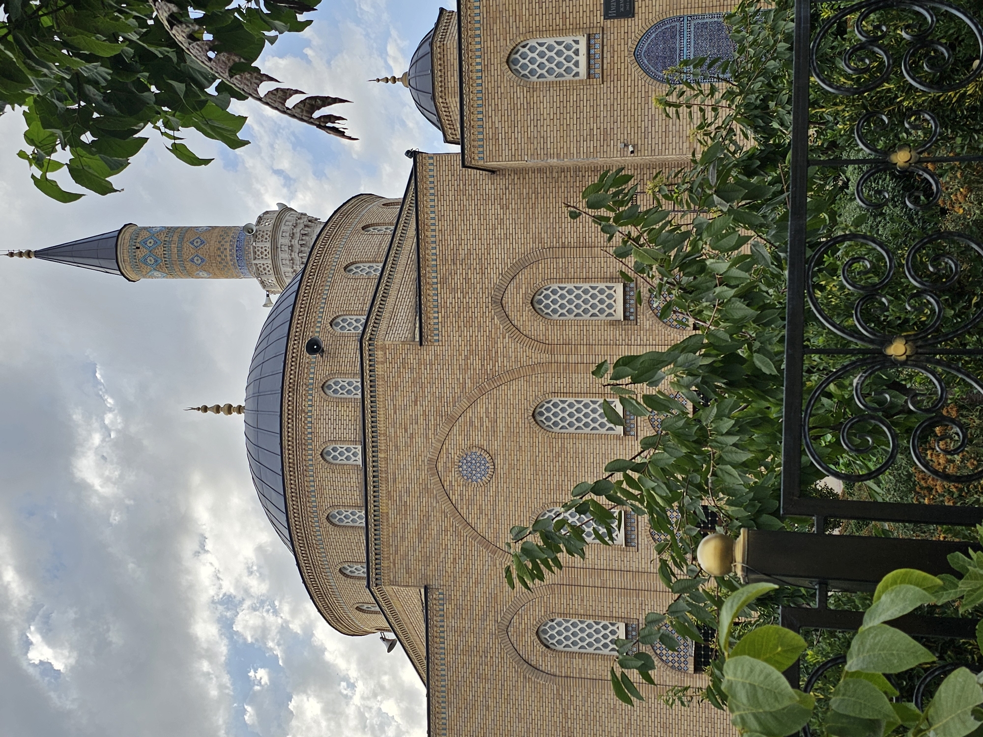 An exterior, eye-level, medium shot captures the Hazrati Shah Mosque in Ozgon, Kyrgyzstan, under a cloudy sky. The mosque's light brown brick facade is dominant, with a large dark dome and a tall, slender minaret topped with a spire. The minaret features intricate blue and light brown geometric patterns. Arched windows with diamond-patterned glass adorn the facade. In the foreground, a black metal fence with decorative scrollwork and a gold finial partially obscures a lush green garden with various plants and trees. The overall scene is peaceful and architectural.