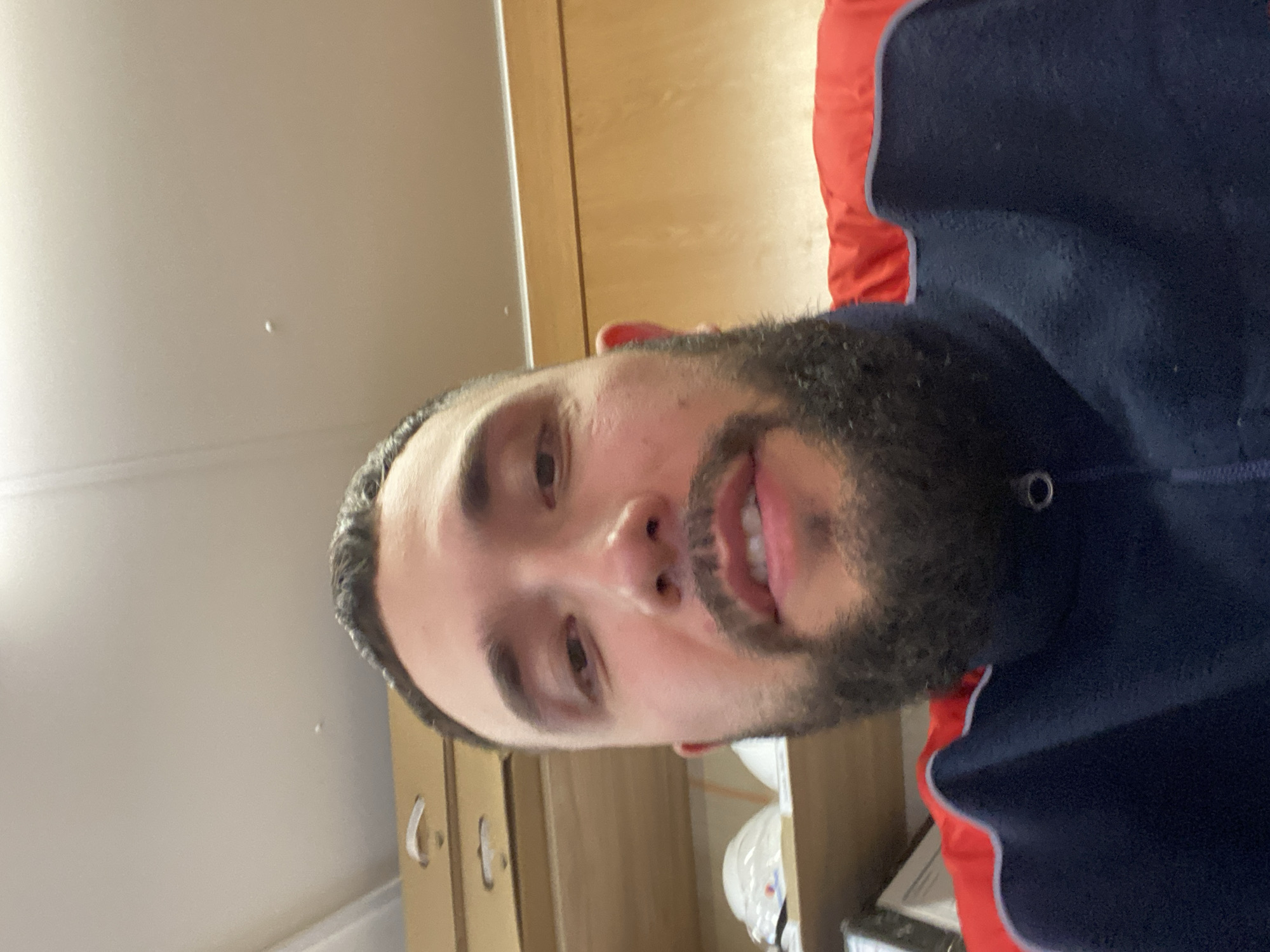 A man with dark hair and a full beard is looking directly at the camera. He is wearing a dark blue and red garment. The background consists of a light-colored wall and wooden cabinets. There is no indication of the city of Luzhitsy, Russia.