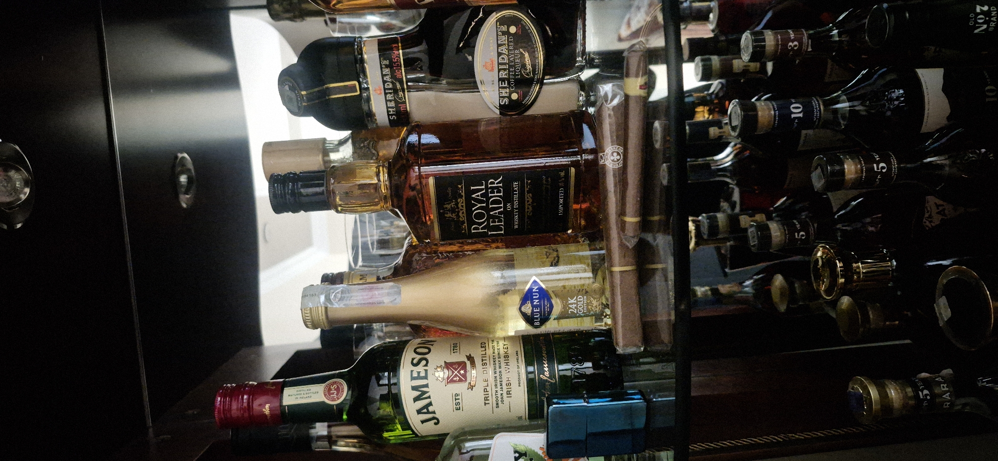 The image shows a liquor cabinet, likely located in Yerevan, Armenia, filled with various alcoholic beverages. Visible bottles include Jameson Irish Whiskey, Royal Leader whiskey, Blue Nun 24K Gold Edition wine, and Sheridan's Coffee Layered Liqueur. There are also bottles of wine, possibly from the Ararat Wine Factory (indicated by the "ArArAt" inscription on a bottle). A pack of cigars is visible among the bottles. The bottles are neatly arranged on shelves within the cabinet. The cabinet door is dark, with metal hardware.