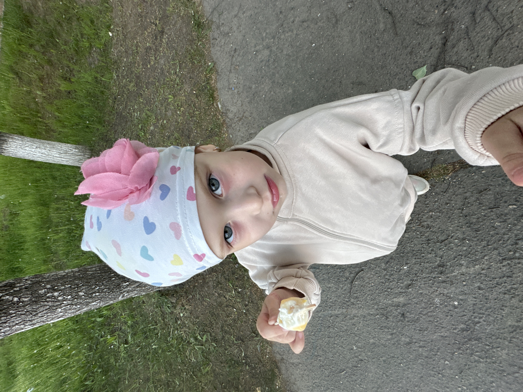 A child is standing outside in what appears to be a park or area with greenery. The child is wearing a light-colored outfit with a hat adorned with a large pink flower and heart pattern. The child is holding an object, possibly a partially eaten ice cream or treat on a stick, and is looking up at the camera. The background includes grass, a tree trunk, and a paved area or path.
The location is Chaikovsky, Russia.