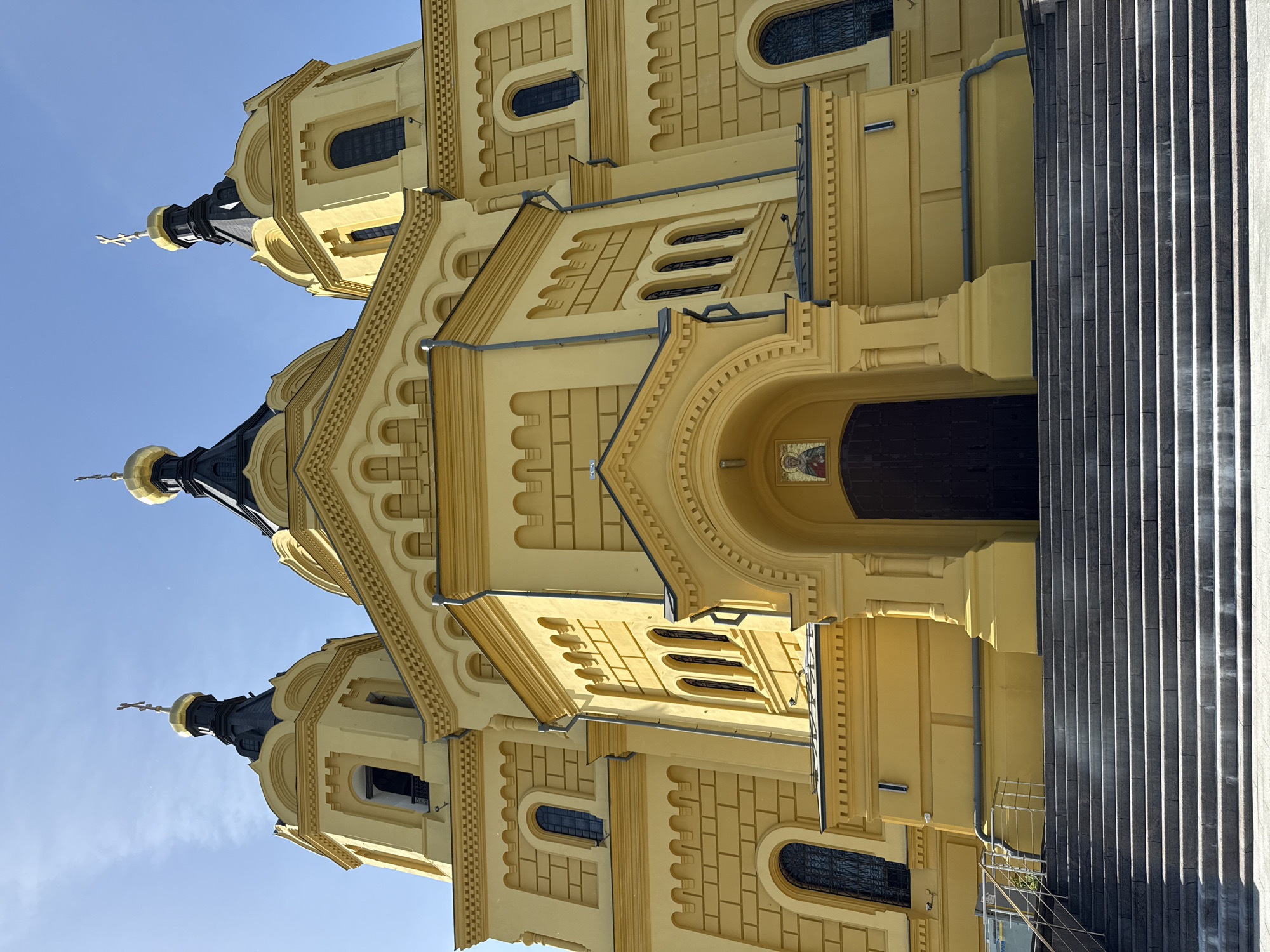 The image shows the Alexander Nevsky Cathedral in Nizhny Novgorod, Russia, from a low-angle perspective. The facade is painted in a yellow tone and has three domed towers topped with golden crosses rising into a blue sky. Architectural details include arched windows, patterned brickwork, and intricate decorative elements around the entrance. To the right of the cathedral is a section of granite stairs, creating a strong vertical contrast with the curved lines of the building. No people are visible in the frame.