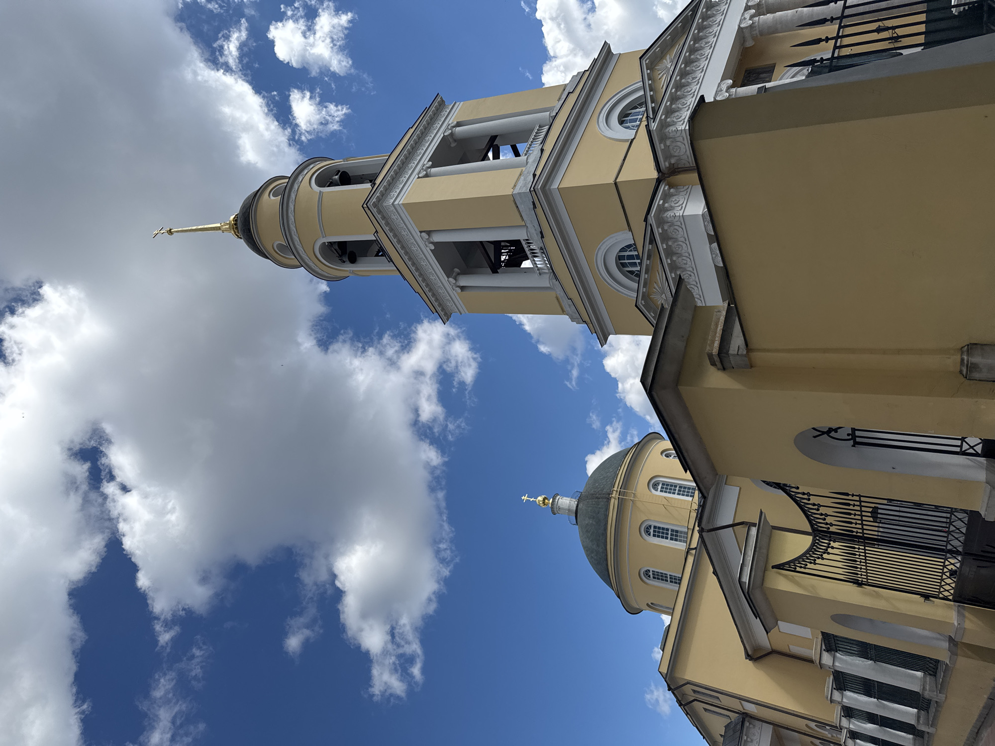 This is a low-angle, exterior, medium shot of the Church of the Ascension of the Lord in Moscow, Russia. The church is painted a pale yellow with white trim and features a prominent bell tower with bells and a gold cross on top. A large, dark green dome with a gold cross is also visible. The sky is blue with fluffy white clouds. A decorative black metal fence is in the foreground. The overall impression is of a sunny day.