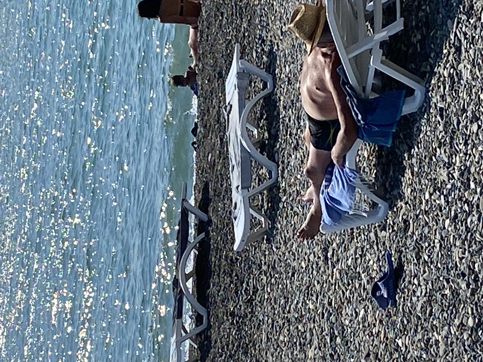 In Chabkwara, Abkhazia, on a pebbly beach, a man wearing a straw hat and black swim trunks lies on a white lounge chair under a bright sun. He is facing away from the camera, with his eyes closed and his arms resting by his side. A blue towel is draped over the lounge chair. Nearby, a single blue sandal rests on the pebbles. The sea, with its surface sparkling from the sunlight, is visible in the background, where a few people can be seen in the water and a child is playing near the shore. Two other empty white lounge chairs are positioned on the beach in the middle ground.
