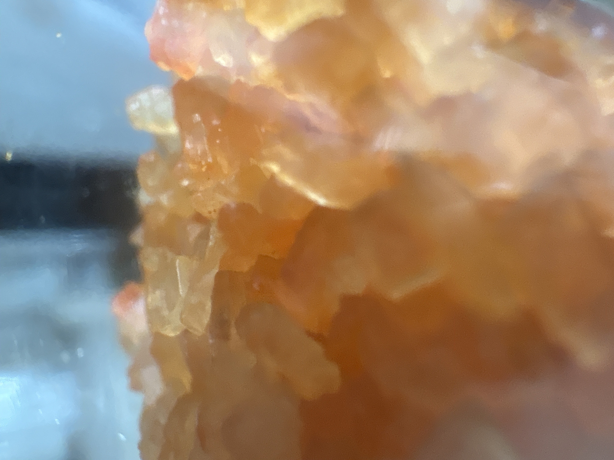 The image shows a close-up view of a translucent, orange-colored crystalline substance. Individual crystal structures are discernible, creating a layered appearance. The left side of the image suggests the substance is contained within a glass or transparent container. The location in the city of Chaikovsky, Russia, is not discernible from the provided image alone. Therefore, no information about the city can be provided.