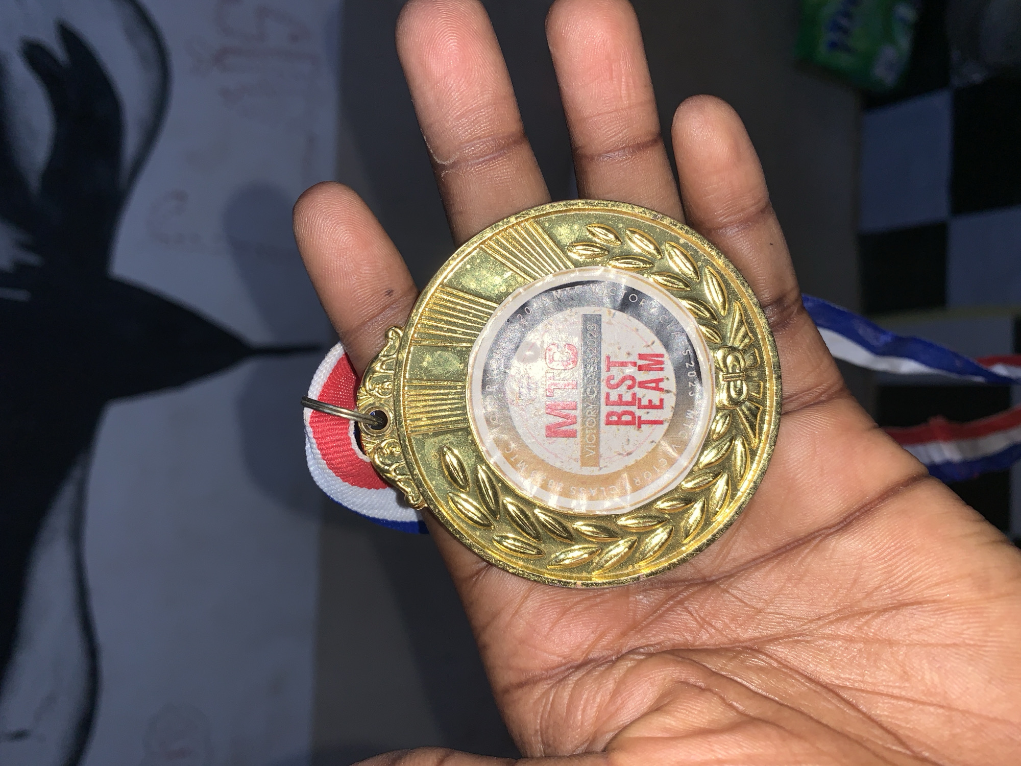 A gold medal is held in the palm of a dark-skinned hand. The medal has an inner circular design with the words "MTC VICTORY CLASS 2023 BEST TEAM" printed on it. The outer ring of the medal has a decorative leaf design. A ribbon with white, red, and blue stripes is attached to the medal. The background is a mix of dark and light areas. The location is Rafin Par, Nigeria.