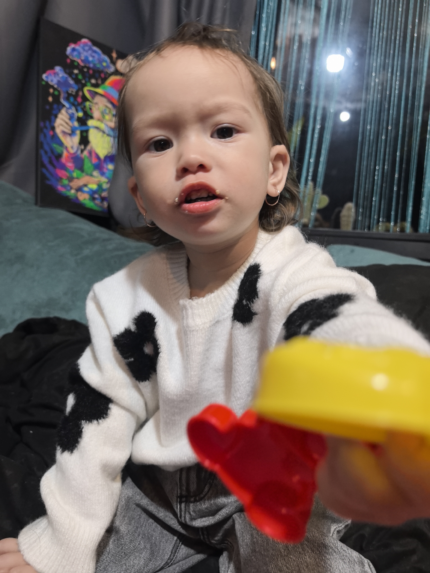 The scene is located in Noviy, Russia. A young child, appearing to be a toddler, is centered in a medium shot, looking directly at the camera. The child has light skin, dark eyes, and light brown hair pulled back, with small gold hoop earrings visible in both ears. The child's mouth is slightly open, revealing teeth and several small, white food particles around the lips. The child is wearing a white knitted sweater with irregular black blotch patterns resembling cowhide, and grey trousers. In the foreground, extending from the child's right hand towards the viewer, is a yellow and red plastic toy. The toy consists of a yellow cylindrical or cup-like top attached to a red handle or base, with some red visible beneath the yellow. The child's left hand is partially visible at the bottom left. The background features a dark green sofa or cushion behind the child. To the left, a vertical rectangular painting is visible, depicting a vibrant, multi-colored figure with a hat against a black background, with blue cloud-like shapes and colorful dots. To the right, a window or glass panel is visible with multiple vertical blue string-like decorations, and two distinct circular light reflections are present in the dark background beyond.