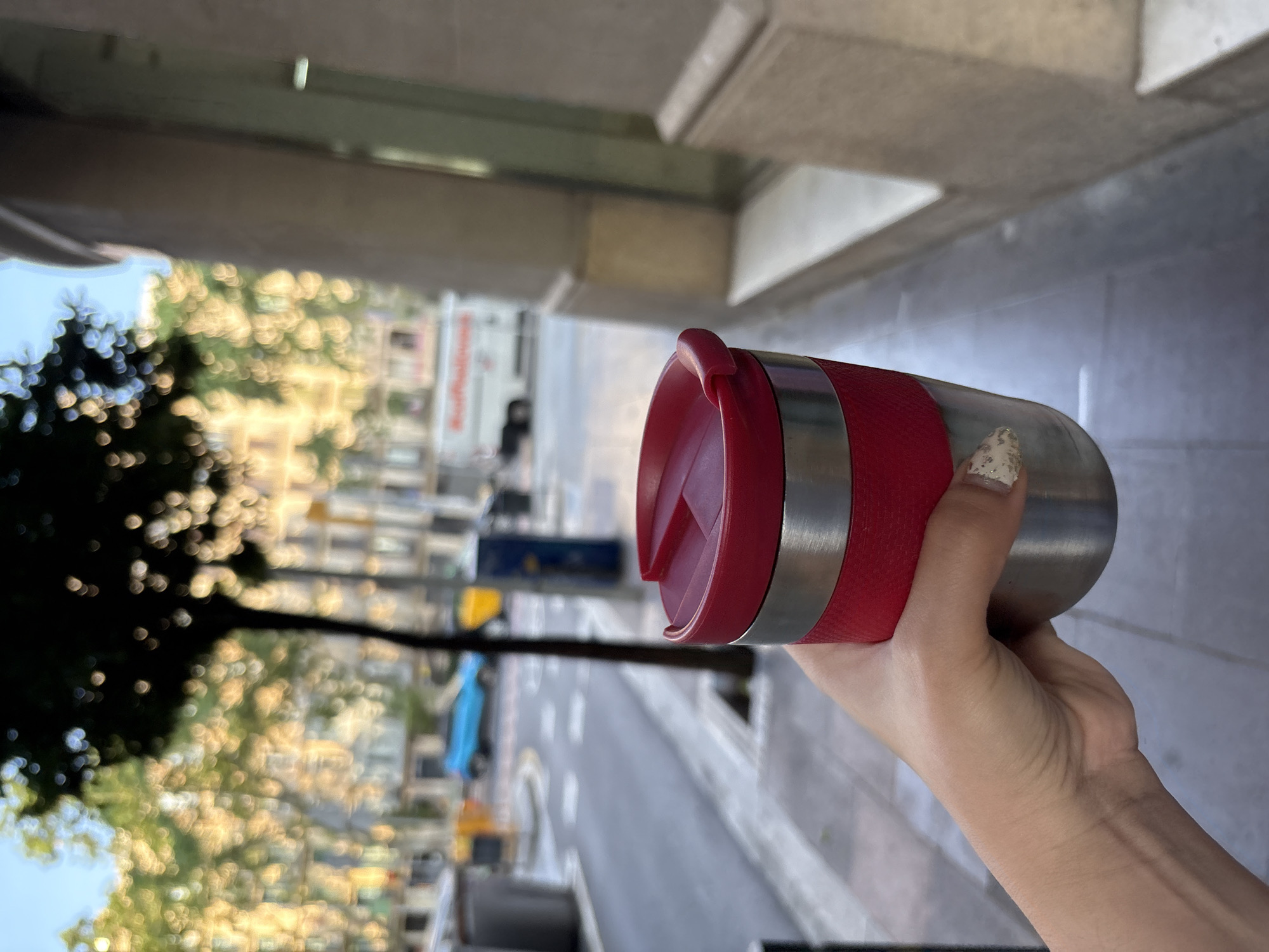 The photo is taken on a city street, likely in Barcelona, Spain, based on the architecture and street layout. A person holds a stainless steel travel mug with a red lid and a red rubber grip in their right hand. The person's hand is fair-skinned with manicured nails. The background shows a typical Barcelona street scene, with buildings, cars, trees, and what appears to be a bus or large vehicle in the distance. The lighting suggests it's daytime. The street is paved and appears to be well-maintained. Above the scene, there is the overhang of a building with a concrete or stone facade. The image seems to be taken from a low angle, looking upwards.