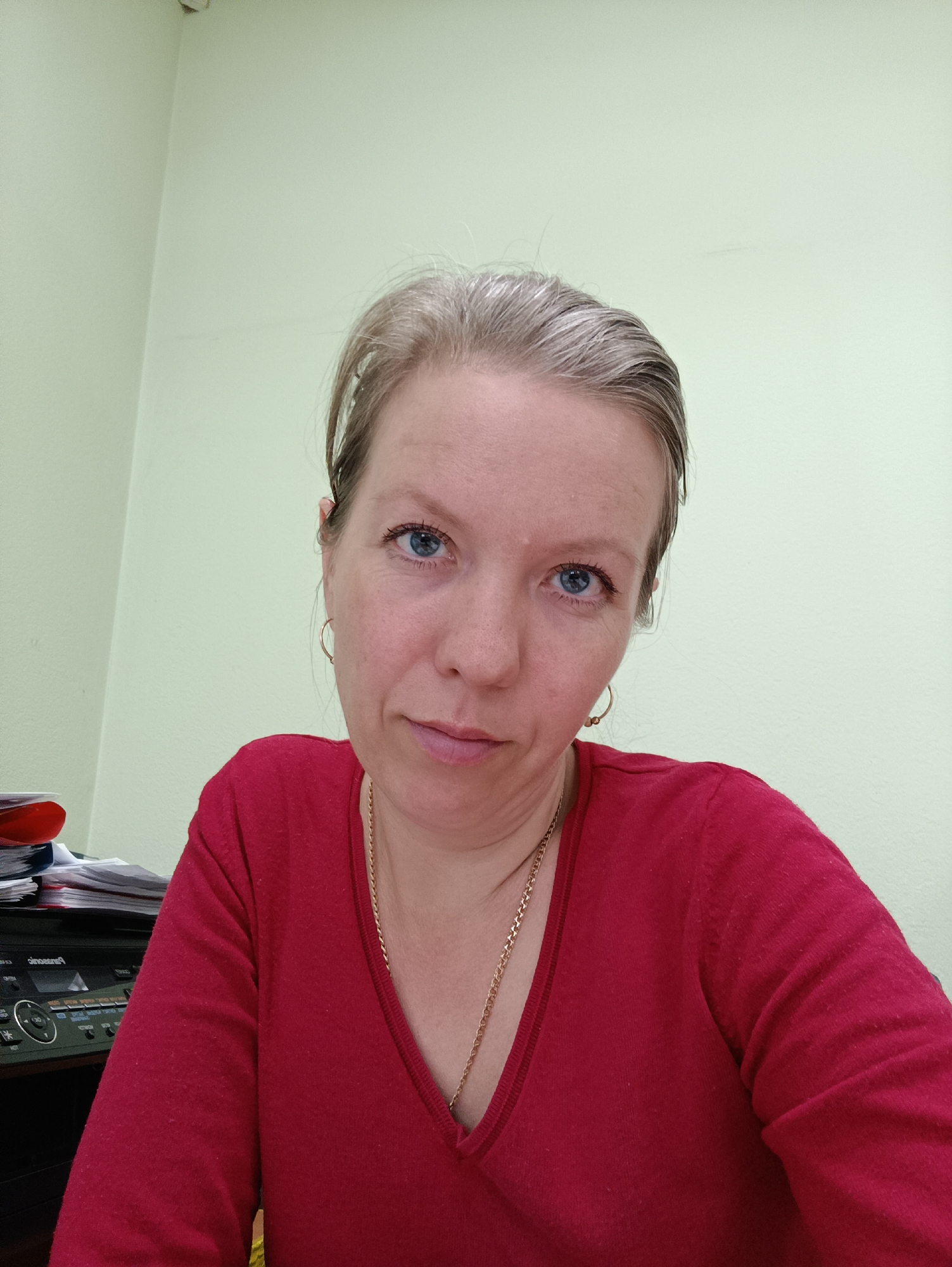 A close-up, full-frame portrait shot of a fair-skinned woman with blonde/graying hair and blue eyes. She is wearing a red V-neck sweater and a gold necklace and hoop earrings. Behind her is a light green wall and a portion of a black printer/scanner with a stack of papers on top. The photo appears to be taken indoors, possibly in an office. The location is Chaikovsky, Russia.