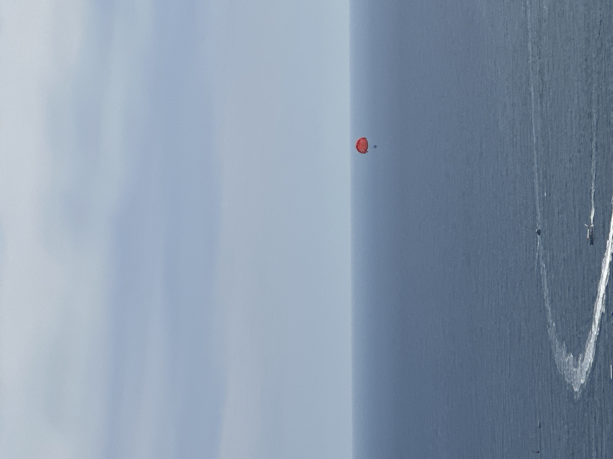 A parasailer in a red canopy is suspended over the vast, deep blue Adriatic Sea, off the coast of Vrela, Montenegro. The parasailer is a small, solitary figure against the immense backdrop of the ocean and the pale, cloudy sky, which occupies the upper two-thirds of the frame. The horizon line is sharp, dividing the water and sky. Closer to the viewer, the water is disturbed by the wakes of several small boats, leaving trails of white foam on the surface. The scene conveys a sense of aerial freedom and the expansive natural beauty of the Montenegrin coastline.