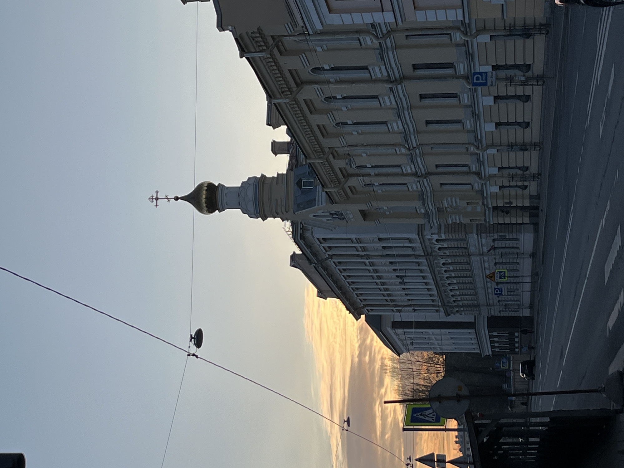 A street scene in Unknown, Unknown, features a tall building with a prominent onion dome topped with a cross, indicating it is likely a religious structure. The building is multi-storied with detailed architectural features, and a parking sign is visible on one of its walls. The street is mostly empty except for a few vehicles. Above the street are overhead wires, and various street signs are visible along the sidewalk. The sky suggests either sunrise or sunset with a soft, golden hue.