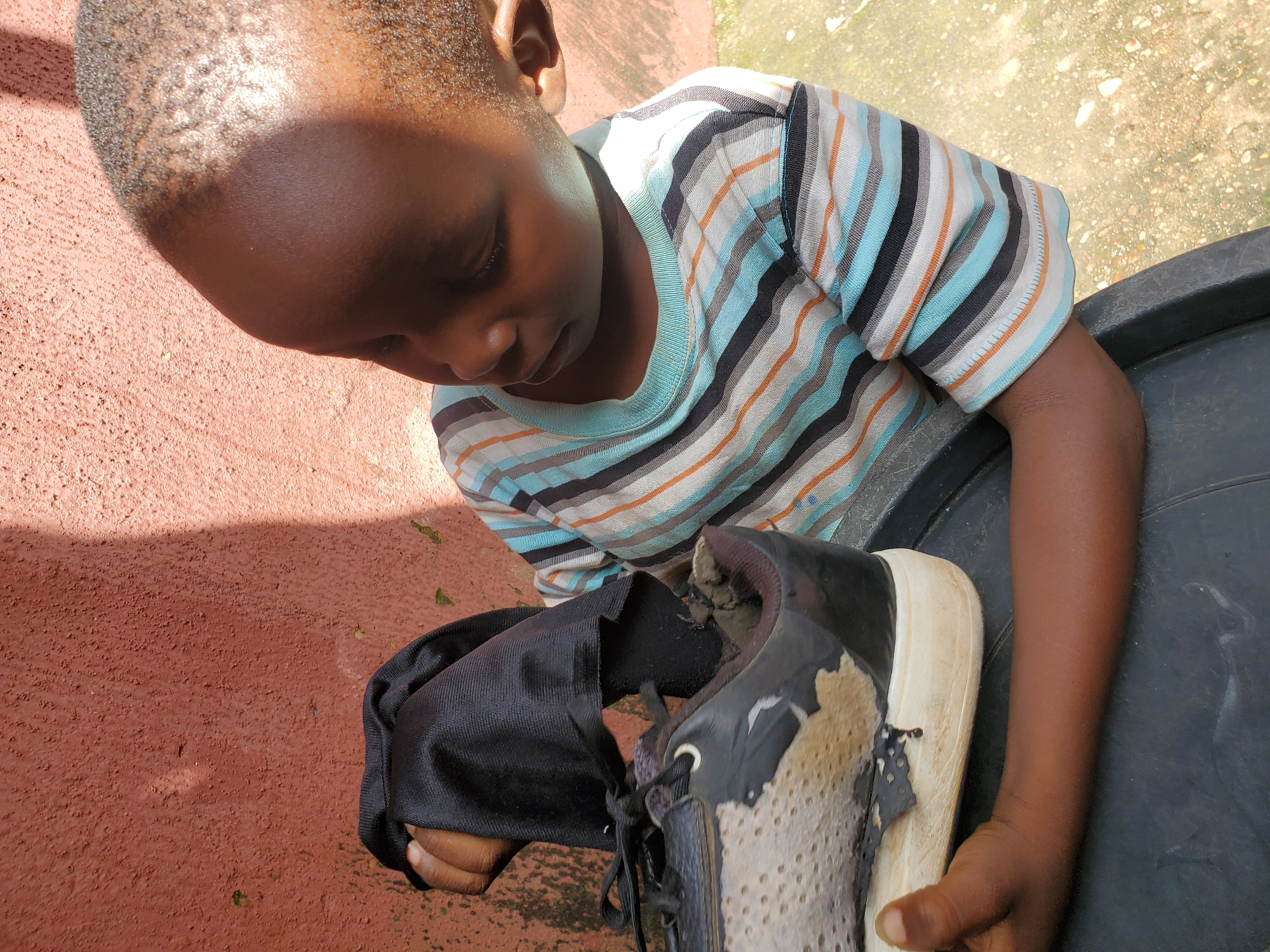 A young Black child, wearing a striped t-shirt with blue, black, orange, and white stripes, is looking down at a worn-out black and white sneaker in their hands. The child's head is bowed, and their gaze is focused on the shoe. One hand is holding the shoe, while the other appears to be on a dark object, possibly a container. The background shows a reddish-brown textured wall on the left and a lighter, speckled surface on the upper right, which could be ground or a pavement. Sunlight casts a shadow on the wall, suggesting it is daytime. The sneaker shows significant wear and tear, with material peeling off and the sole discolored. The scene appears to be outdoors, possibly in a residential area given the wall and ground texture. The overall impression is one of a child examining a damaged item. The location is identified as Jos, Nigeria. There is no visible text in the image.